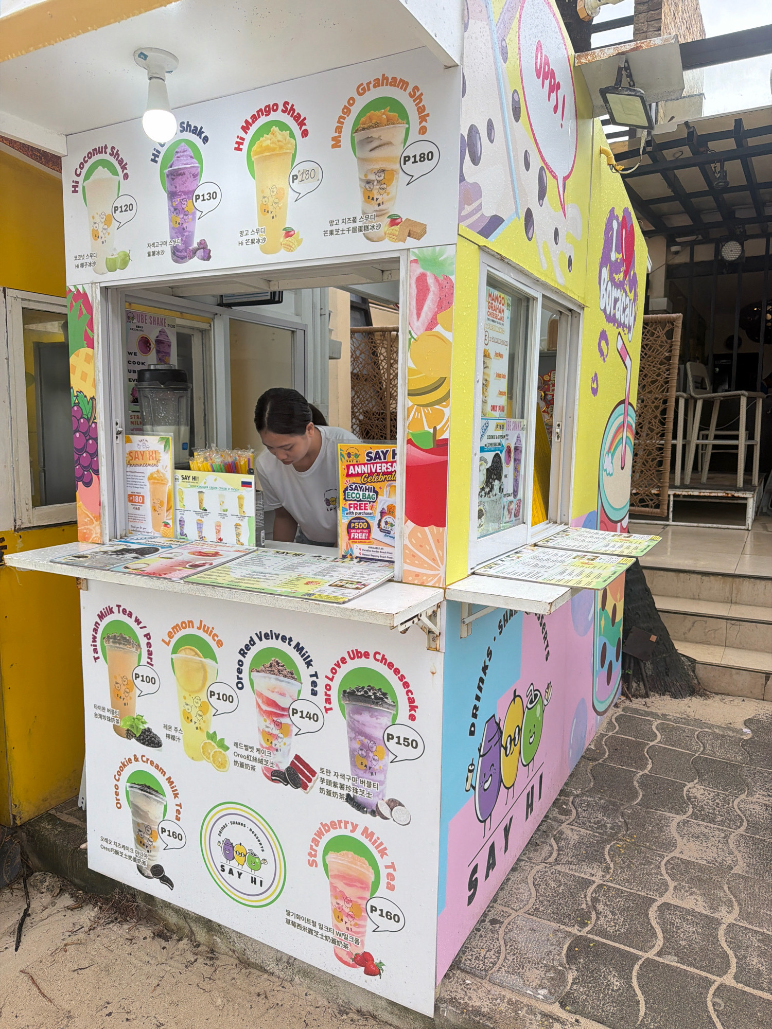 An outdoor beverage stall, branded "SAY HI," is depicted in Malay, Philippines. The stall is vibrant and colorful, adorned with cartoonish illustrations of fruits and drinks. It features a menu board showcasing various shakes and milk teas, with prices indicated in Philippine Pesos. A young woman, presumed to be an employee, is seen behind the counter, attending to customers or preparing orders. The stall is situated on a sandy and paved ground, suggesting it's a beachfront or tourist area. The architecture of the background suggests a tropical, possibly resort-like setting, common in the Philippines. The overall scene captures a casual, commercial environment focused on serving refreshing beverages.