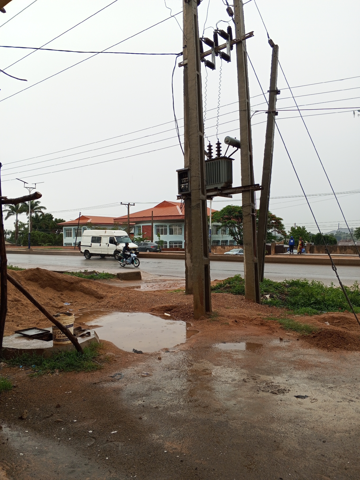The image depicts a street scene in Unknown, Unknown. In the foreground, the ground is wet and muddy, with puddles and debris scattered around. Construction materials, including a bucket and sand, are present. Three concrete electrical poles stand prominently, with a transformer attached to one. Power lines crisscross the sky. In the background, a white van and a person on a motorcycle are crossing the street. Other vehicles and pedestrians are also visible. Buildings with red roofs are located further back across the road. The sky is overcast and gray.