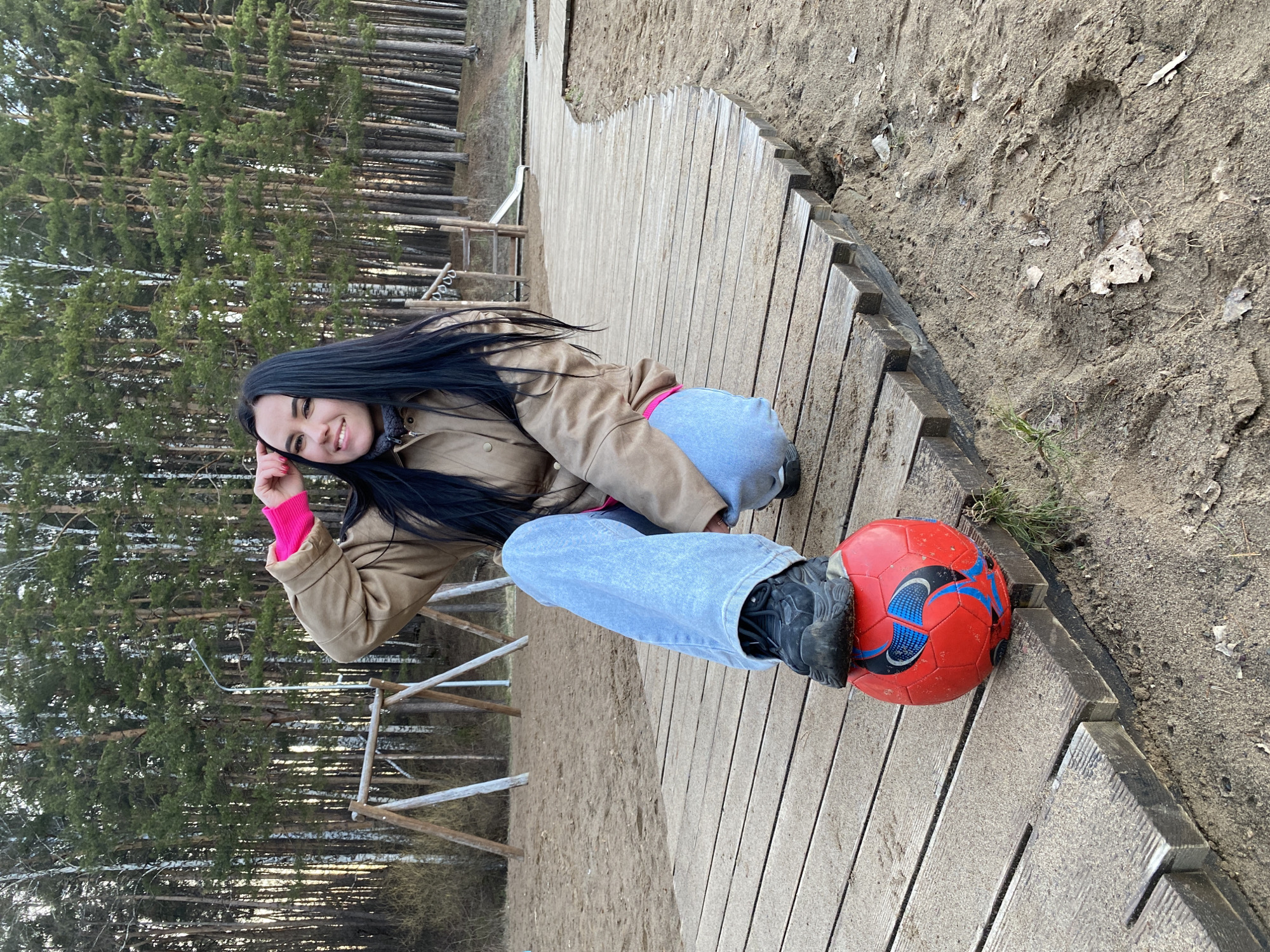 A young woman is captured in a full-body shot, kneeling or crouching on a rustic wooden boardwalk that curves through a sandy, outdoor environment. She has long, dark hair and is smiling warmly at the viewer, with her right hand gently touching her chin or cheek. She is dressed in a light brown or beige jacket, a bright pink long-sleeved top visible underneath, and light blue jeans. Her left foot, clad in a black athletic shoe, rests on a red soccer ball with black and blue accents.
The scene is set outdoors in what appears to be a natural park or forested area. Behind the woman, tall, slender pine trees dominate the background, suggesting a dense forest. Some bare birch trees are also visible among the pines. The ground adjacent to the wooden path is sandy or dirt, with sparse vegetation and fallen leaves. In the mid-ground, simple wooden structures, possibly part of a playground or natural trail feature, are visible. The lighting suggests it is daytime, possibly overcast or late afternoon, as there are no harsh shadows. The overall mood conveyed by the woman's smile is cheerful and relaxed. There is no discernible text visible in the image. This scene is consistent with outdoor recreational areas that might be found in Chaikovsky, Russia.