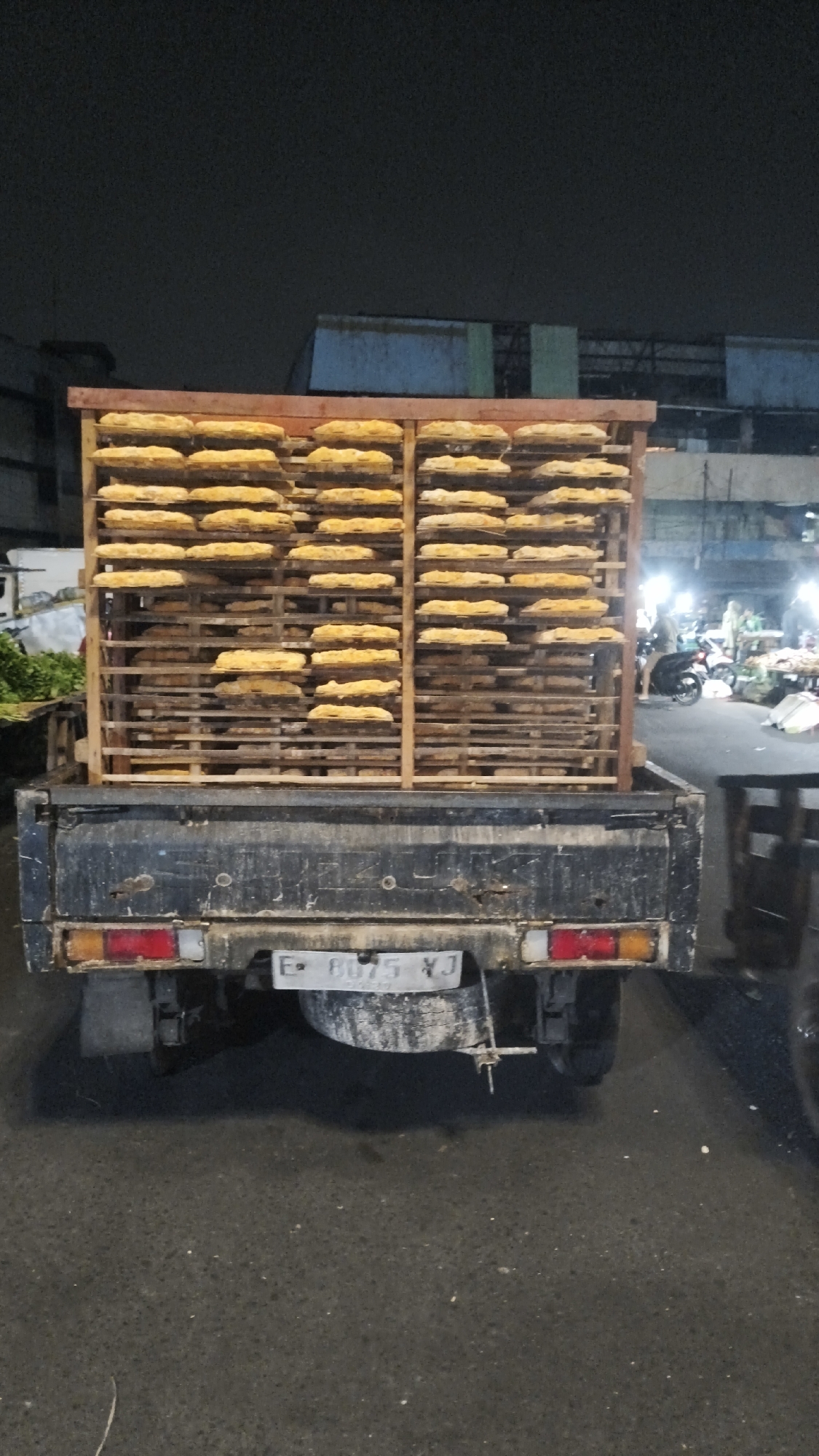 A flatbed truck, parked on a paved surface at night, is loaded with a wooden rack filled with many flat, rectangular, yellow objects stacked on shelves. The truck's license plate is visible and reads "E 8075 YJ". The background shows buildings, streetlights, and other vehicles, suggesting an urban or market environment in South Jakarta, Indonesia. There are no people clearly visible in the foreground.