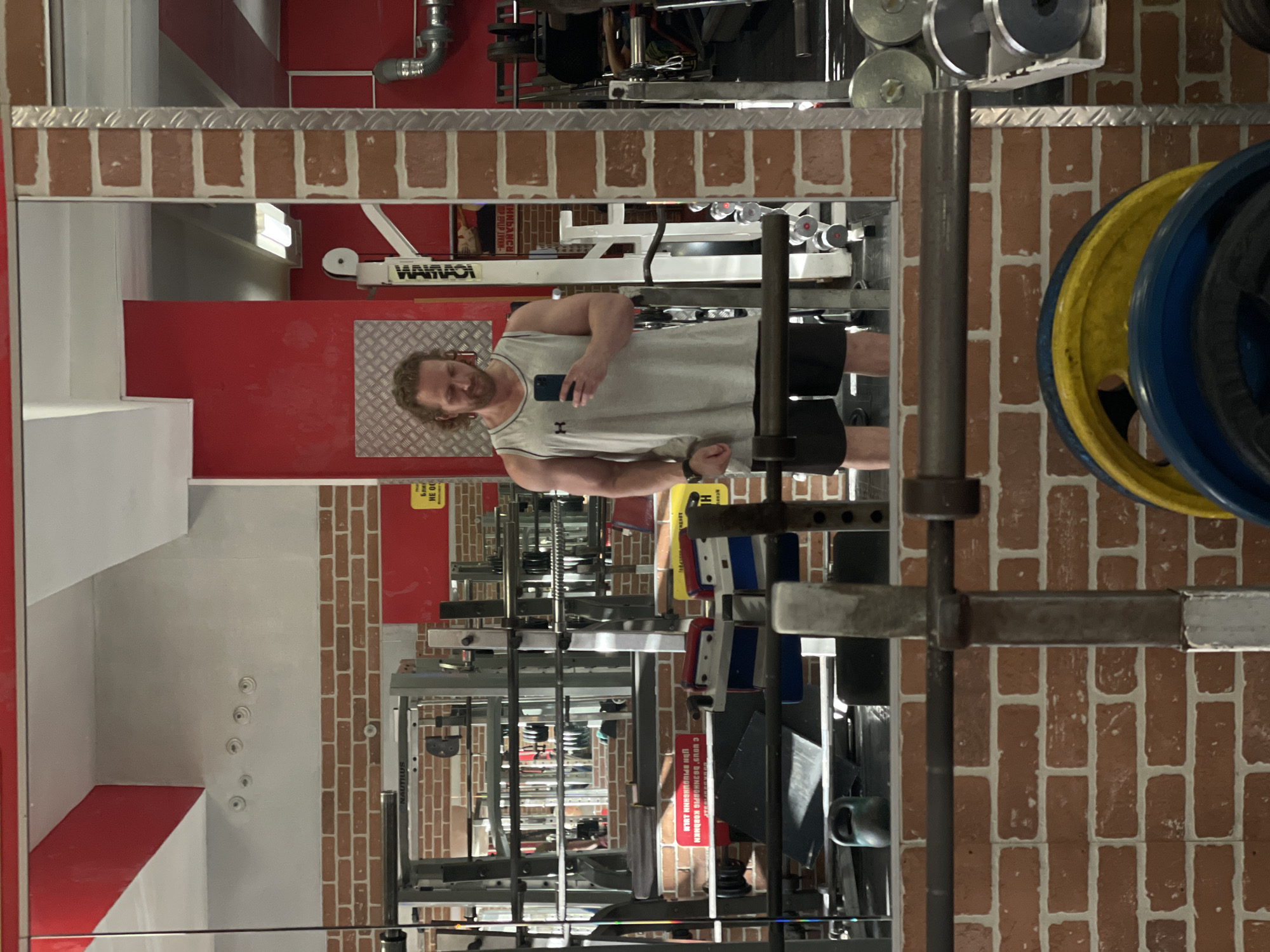 A man with curly blonde hair is taking a mirror selfie in a gym. He is wearing a light gray tank top and black shorts. In the background, there are various gym equipment, including weight racks, barbells, and weight plates. The walls are covered in red brick. The gym appears to be located in Saint Petersburg, Russia, as indicated by a sign on the wall with Russian text.