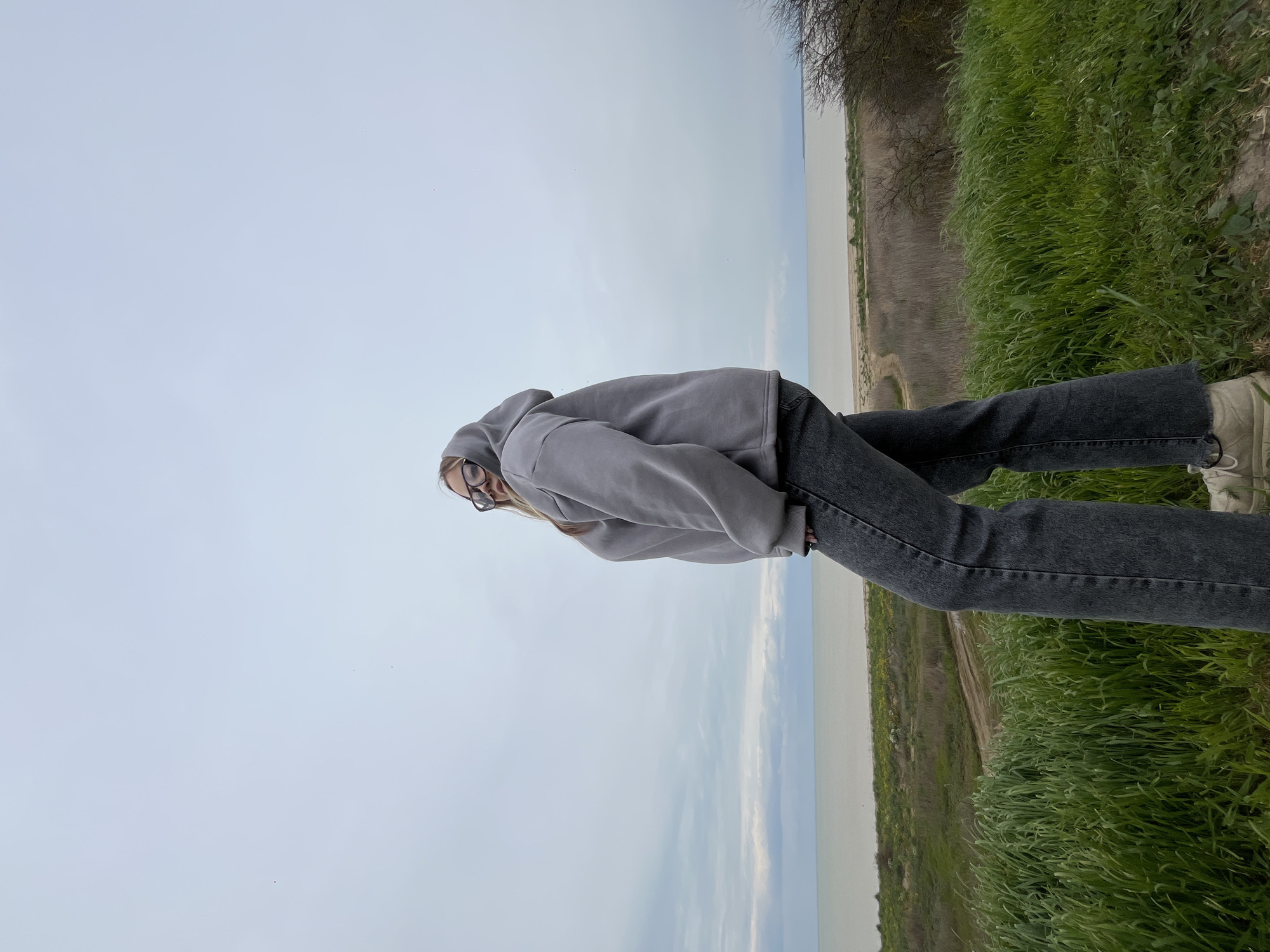 A person wearing glasses, a gray hoodie, and dark jeans is standing on a grassy bank overlooking a body of water. The person is positioned in the center of the frame, with the water and sky occupying the left side and the grassy bank extending to the right. The location appears to be outdoors, possibly near a lake or river. The sky is overcast. There are no other people visible in the frame. The specific location in Port-Katon, Russia is not discernable.