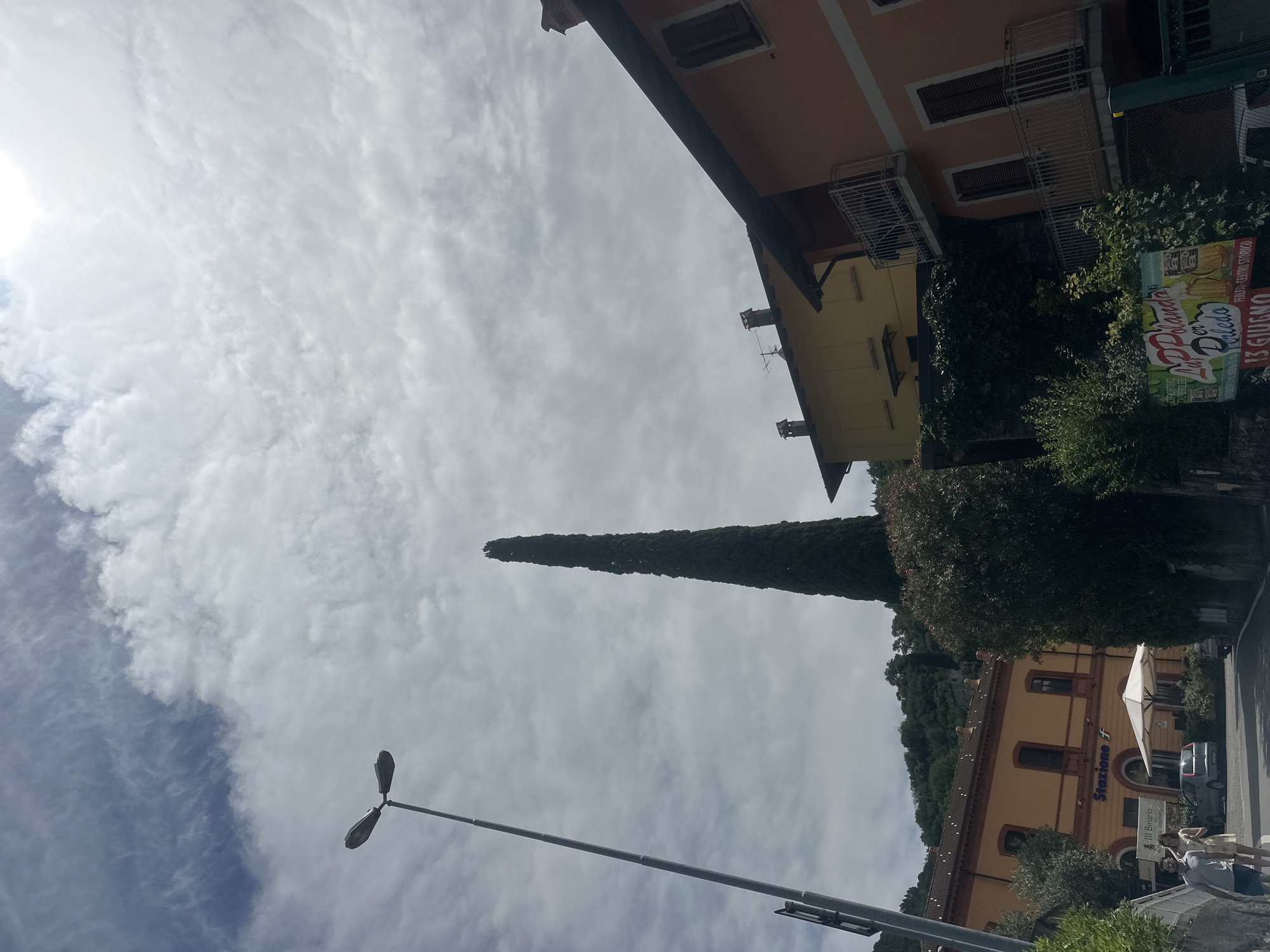 The image shows a street scene in Gittana, Italy. On the left, a tall lamppost stands against a cloudy sky. Behind it, a yellow building with arched windows displays the word "Stazione f," indicating it is a train station. A few people are walking on the sidewalk in front of the station. Further to the right, a very tall, slender cypress tree dominates the scene. Behind the tree, two multi-story buildings are visible: a yellow one with visible chimneys and a reddish-orange one with balconies and shutters. A sign with an advertisement is visible on the lower right side of the image, featuring images of beer and text. The overall setting appears to be a town, possibly with a Mediterranean feel, given the architecture and the lush greenery in the background.
