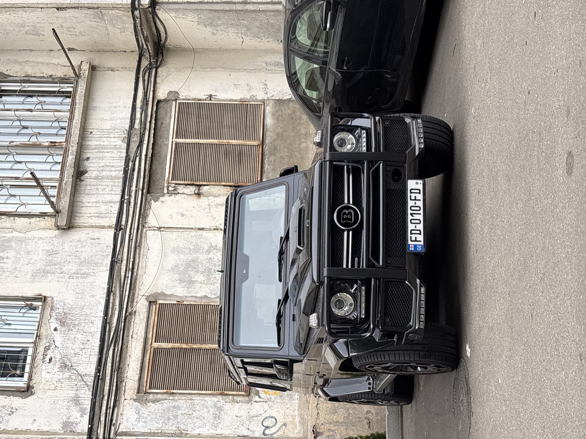 A black Brabus-tuned Mercedes-Benz G-Class SUV is prominently featured, parked on a paved street next to a weathered building. The image is oriented vertically, showing the vehicle’s front from a slightly elevated angle. The SUV is sleek and dark, with a distinctive grille bearing the circular 'B' logo of Brabus and "BRABUS" written below it. Its headlights are round, featuring multiple LED elements, and large, custom multi-spoke wheels are visible. The license plate, "GE FD-010-FD," is clearly visible on the front bumper, indicating the vehicle is registered in Georgia, which aligns with the given location context of Tbilisi.
Behind the Brabus, another black car, possibly a sedan, is partially visible, parked closely. The setting is an urban environment, with a light-colored, aged building facade directly adjacent to the street. The building shows signs of wear, including visible cracks, peeling paint or plaster, and dark stains. Several black electrical cables are haphazardly routed vertically down the building's exterior. Two distinct windows with decorative metal security grilles are visible on the building, along with two rectangular vents covered by vertical metal slats. The ground is a light grey paved surface, likely asphalt, and a circular manhole cover is partially visible near the front passenger wheel of the SUV. The lighting suggests it is daytime, possibly overcast, given the soft shadows. No people are visible in the scene. The overall impression is a street parking scenario in an older, lived-in part of a city.
