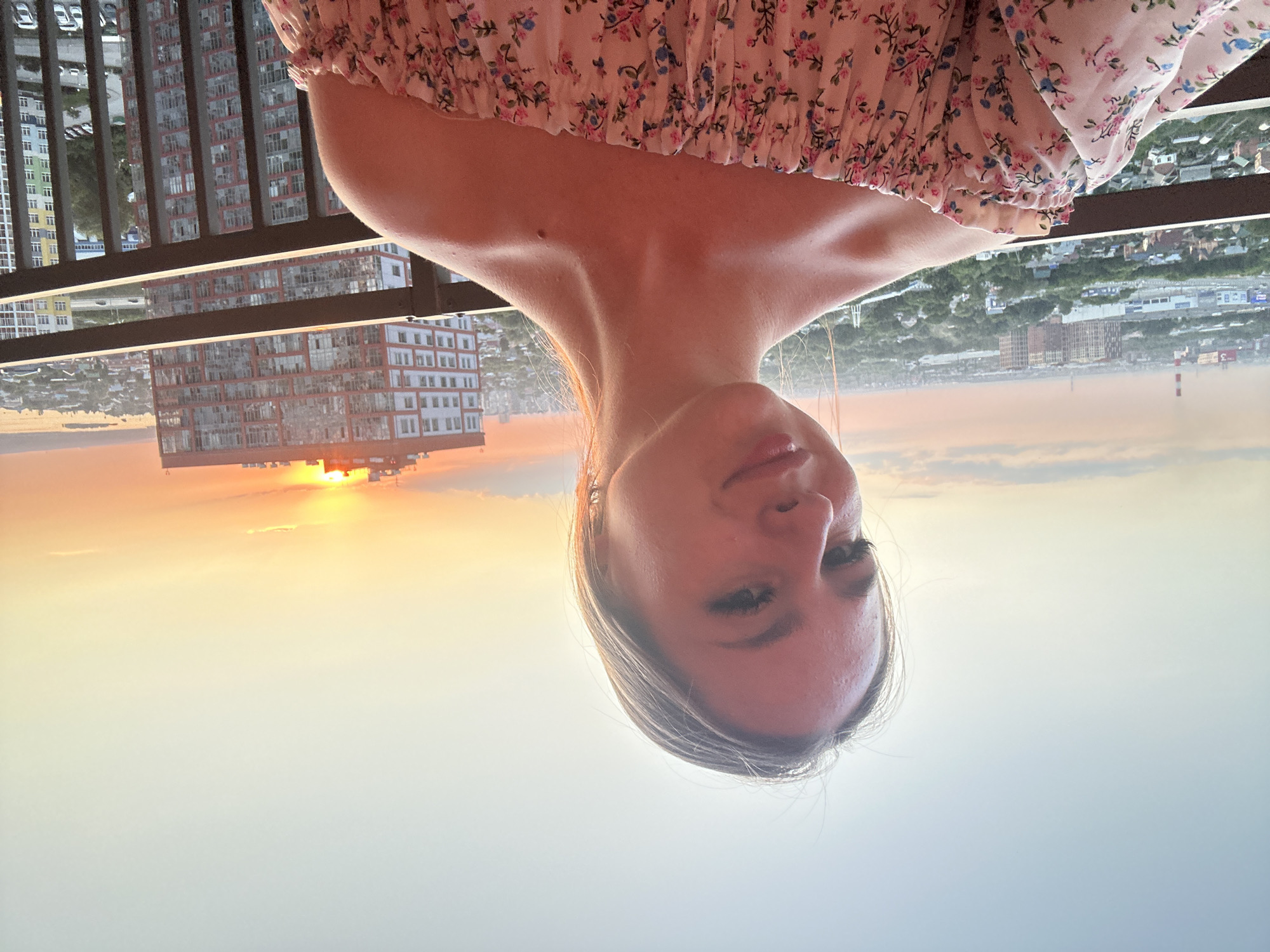 A young woman with blonde hair takes a selfie on a balcony overlooking a cityscape at sunset. She is wearing an off-the-shoulder floral print top. In the background, the sun is setting behind a tall, modern building, casting a warm glow over the city. The balcony railing is visible in the foreground.