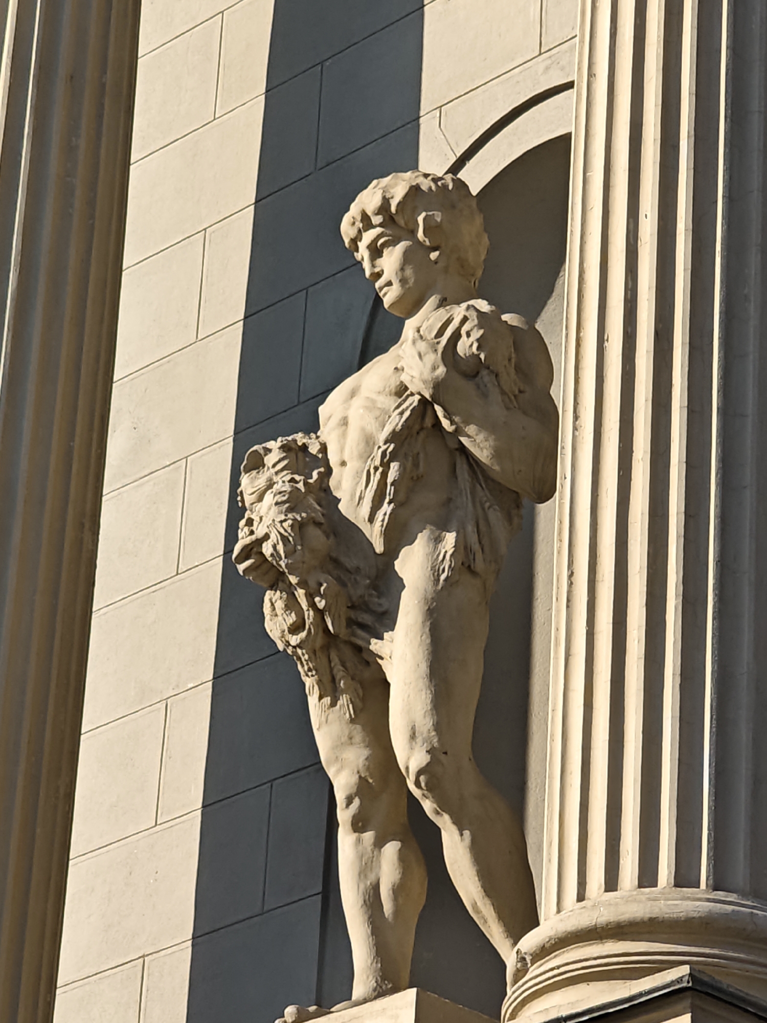 The image is a medium shot of a stone sculpture of a young man holding a lion's pelt in front of a building facade. The sculpture is positioned between two fluted columns. The building facade is made of large, rectangular stone blocks. Sunlight casts a shadow diagonally across the facade. The sculpture is detailed, showing the musculature of the young man and the texture of the lion's pelt. The overall scene suggests an outdoor architectural setting, likely in a city. The specific location in Moscow, Russia, is not discernible from the image itself.
