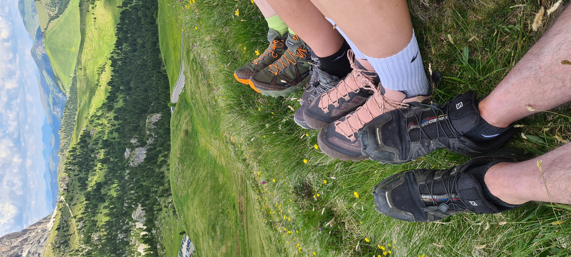 The image shows a close-up view of four people's legs and hiking boots resting on a grassy hillside. The background features a sweeping landscape of green rolling hills dotted with pine trees, a winding road, and distant mountains under a cloudy sky. The location is identified as Sëlva - Wolkenstein - Selva di Val Gardena, Italy, a region known for its mountainous terrain and hiking trails. The people are wearing various hiking boots, including black Salomon boots, pink and black boots, and green and orange boots. Some are wearing socks, one pair being white and another lime green. The grass is green and interspersed with small yellow wildflowers. The scene suggests a break during a hike, with individuals relaxing and enjoying the scenic view.