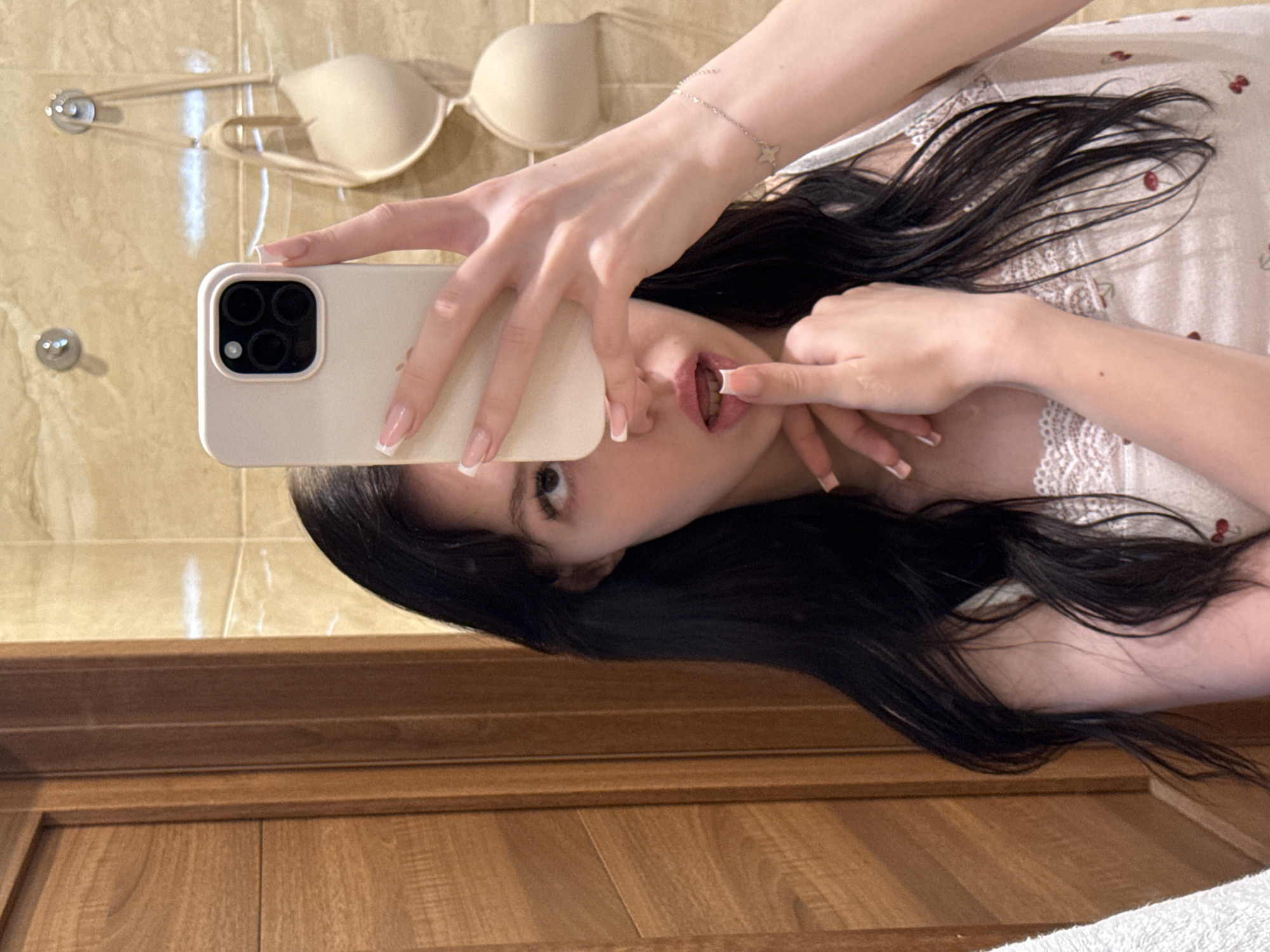A young woman with long, dark hair takes a mirror selfie in a bathroom with tiled walls and wooden accents. She is holding a smartphone in her right hand, partially obscuring her face. She is wearing a light-colored top with lace trim and a cherry print. A beige bra hangs from a hook on the wall behind her. Her left hand is raised to her mouth, with her index finger touching her lips. A delicate bracelet with a four-petal flower charm is visible on her left wrist. The scene is likely a residential bathroom in the city of Khimki, Russia.