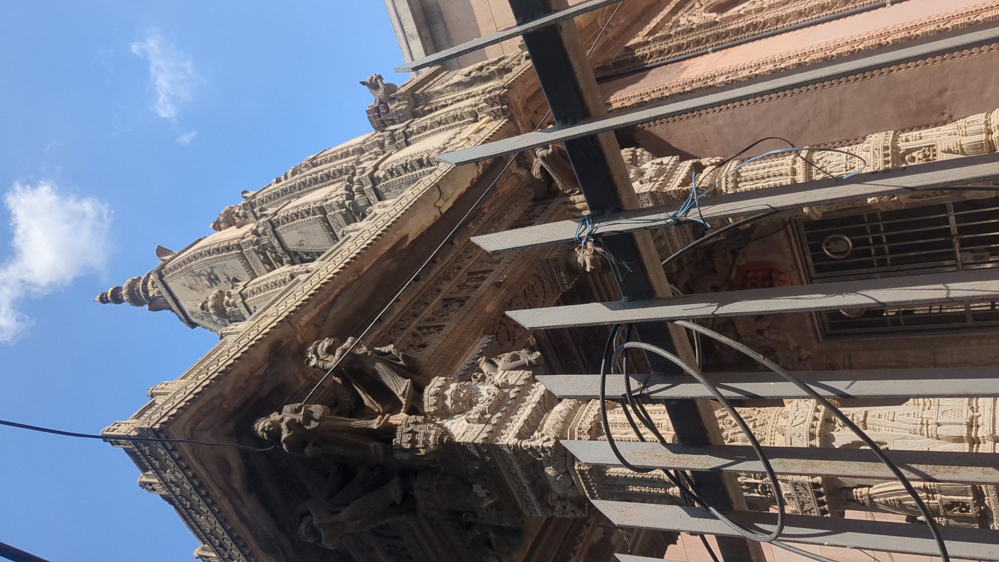 A low-angle, vertical shot showcases the ornate facade of a Hindu temple in Varanasi, India, under a bright blue sky with scattered clouds. The temple exhibits intricate carvings and architectural details characteristic of Indian temple design, including tiered spires, decorative friezes, and sculpted figures adorning its exterior. A metal fence with vertical bars and horizontal supports runs across the foreground, partially obscuring the lower portion of the temple. Electrical wires are visible, strung across the scene and looped around some of the fence structures. The overall impression is one of ancient architecture set against a vibrant sky, with modern infrastructure elements present.