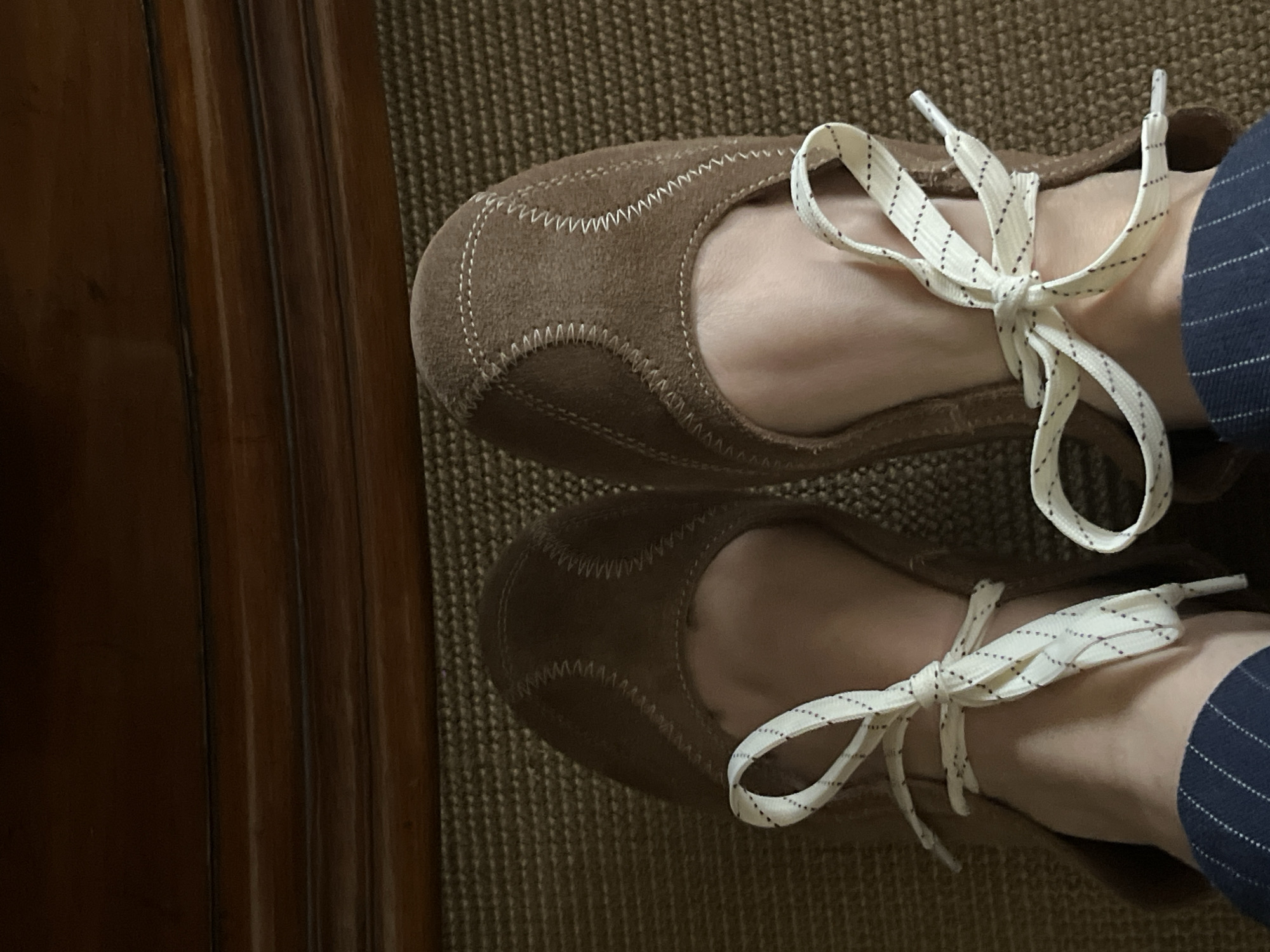 A top-down shot shows a person's feet wearing brown suede flats with white, dotted shoelaces tied in bows. The person is also wearing a dark blue pinstripe pant leg. The background is a brown textured rug or carpet. To the left of the shoes, a wooden door or cabinet edge is visible. The scene does not suggest a location in Mexico City or provide any indication of being in that location.