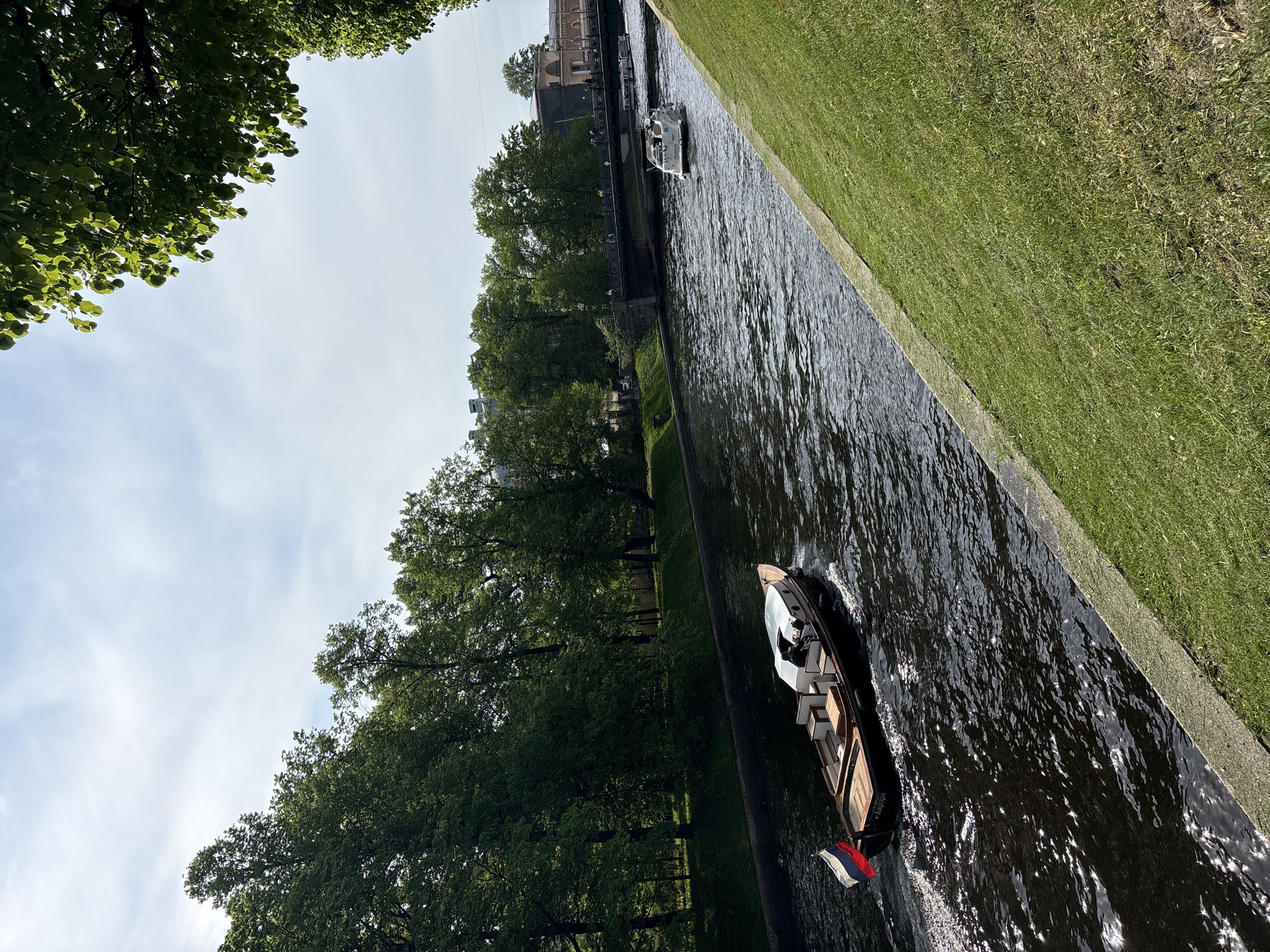 The image shows a canal in Saint Petersburg, Russia, likely during daylight hours. A motorized boat with a flag that appears to be the Russian tricolor is moving along the canal. There is at least one person visible on the boat. Along the canal bank, there is a strip of green grass. Across the canal, mature trees line the other bank. In the background, there's a glimpse of buildings. A smaller boat is visible further down the canal. The sky is partly cloudy.