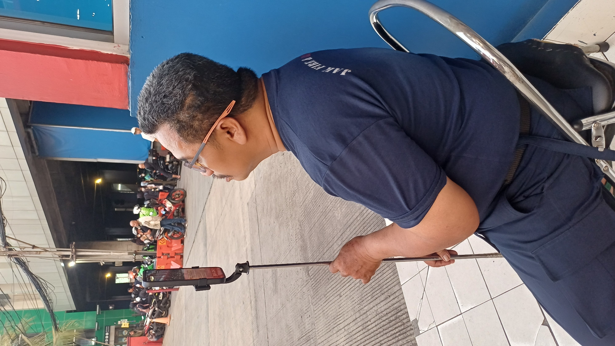A man wearing a navy blue shirt and pants is seated on a silver folding chair outdoors. He is holding a selfie stick with a smartphone attached. The background shows a street scene with several motorcycles and people, likely waiting for rides, in front of an elevated structure that appears to be an overpass or bridge. Blue and red walls are visible to the right of the man. The ground is paved with concrete and white tiles. The overall setting suggests a busy urban environment in South Jakarta, Indonesia.
