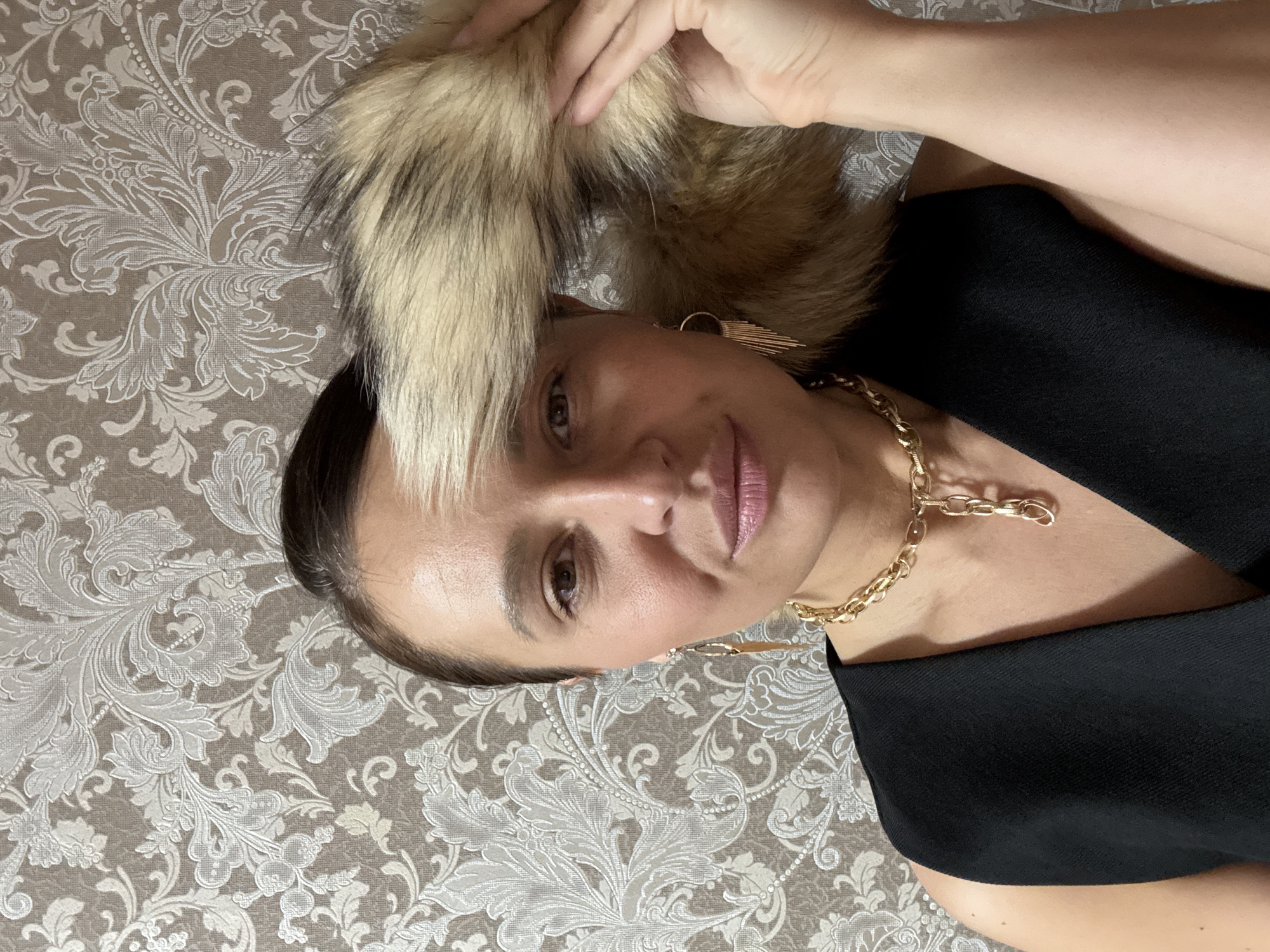 A close-up, angled indoor portrait features a woman with dark hair pulled back, looking towards the camera with a neutral to subtly smiling expression. Her lips are a light pink. A large, fluffy, light-colored furry object, possibly an accessory like a fur collar or trim with blonde and darker tones, is positioned near her head and partially touched by her visible left hand. She wears a black top or dress, adorned with a chunky gold chain necklace and long, geometric gold earrings. The background consists of patterned wallpaper with intricate light beige floral and damask motifs on a muted beige-grey base. The lighting suggests an indoor setting, likely artificial. The image provides no specific visual cues to its location, though it is contextualized as Chaikovsky, Russia. There is no visible text.