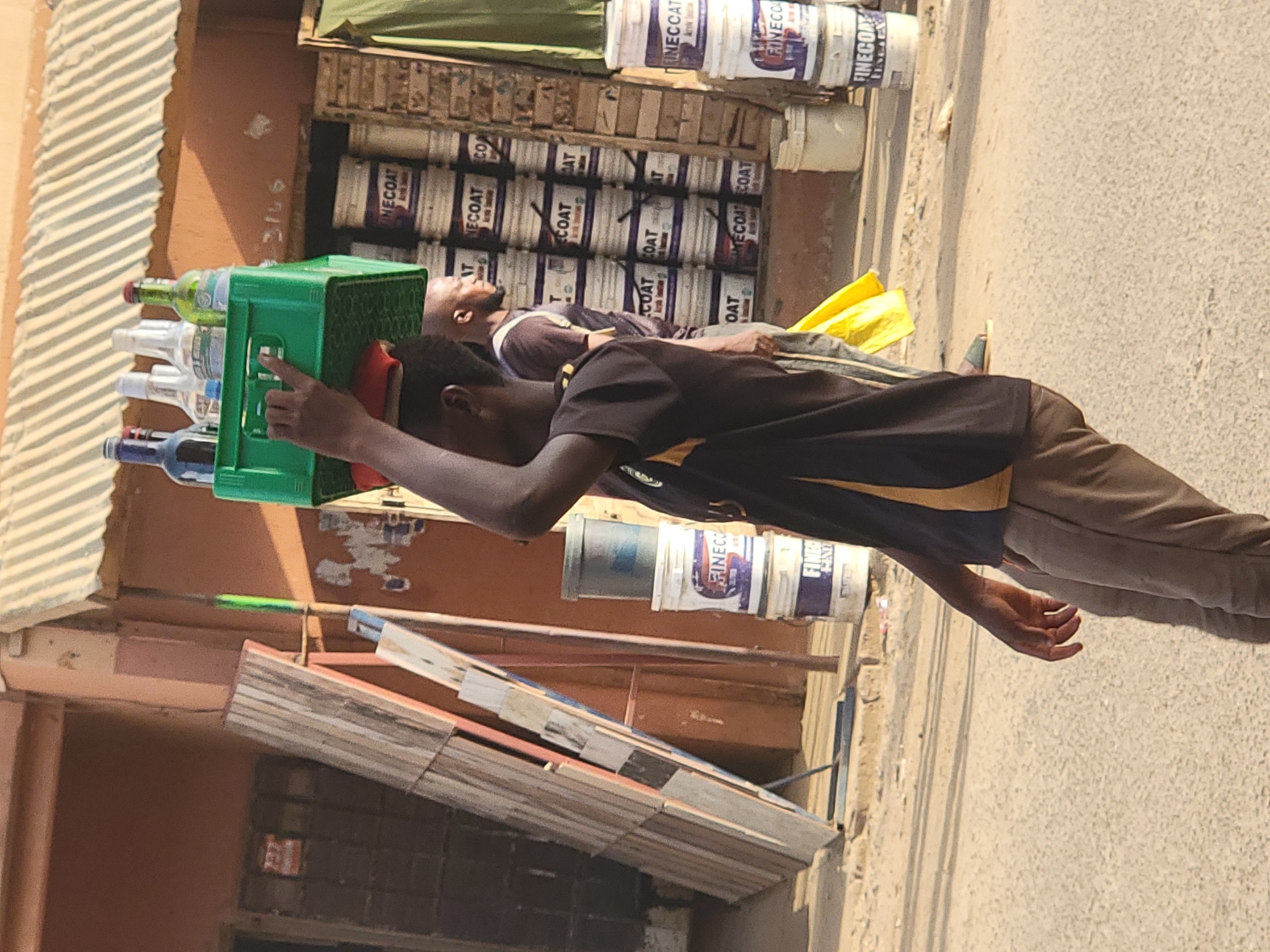 In Maiburiji, Nigeria, a man carries a green crate filled with bottled beverages balanced on his head. He is dressed in a dark t-shirt with a yellow stripe on the shoulders and brown trousers. He appears to be on a street. Behind him, another man is lying on a pile of paint cans, and stacks of paint cans are visible in the background. The environment includes a storefront and an adjacent building.
