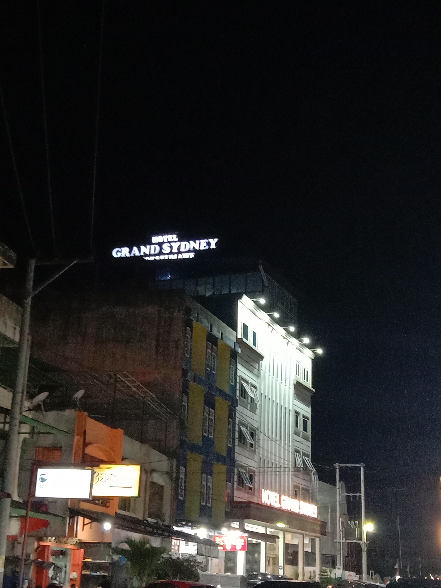At night, the Hotel Grand Sydney in Lhokseumawe, Indonesia stands illuminated. The hotel's sign, "HOTEL GRAND SYDNEY," is brightly lit atop the building. The hotel's facade has sections colored white, yellow, and blue, with numerous windows visible. Several lights illuminate the front of the building at ground level. Utility poles and wires are visible in the foreground. Street signs glow with artificial light. The overall scene is urban and lit by electric lights. The sky is dark and clear. There are plants.