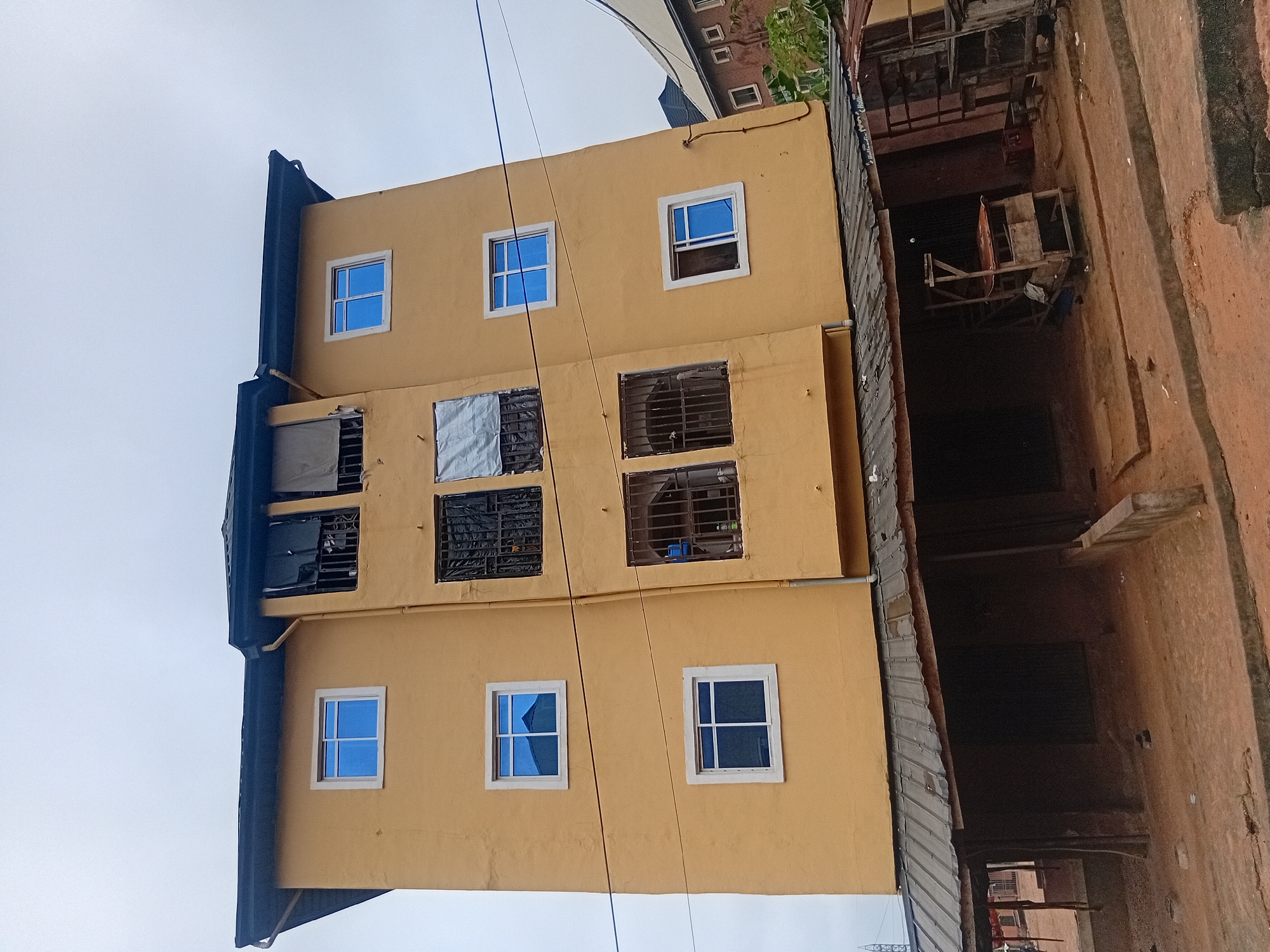 The image depicts a three-story building with a yellow facade in Nkpor, Nigeria. The building has a dark, corrugated metal roof. There are several windows visible on the upper floors, most of which are rectangular and filled with blue glass. Some windows on the second floor appear to be barred or covered. The lower level of the building features an open-air structure with a corrugated metal roof supported by wooden poles, suggesting it may be a shop or market stall area. The ground in front of the building is dirt and appears somewhat uneven. In the foreground, there are concrete barriers or steps. To the right of the yellow building, a portion of another building with a darker roof and windows is visible, along with some greenery. The sky is overcast and a pale gray.