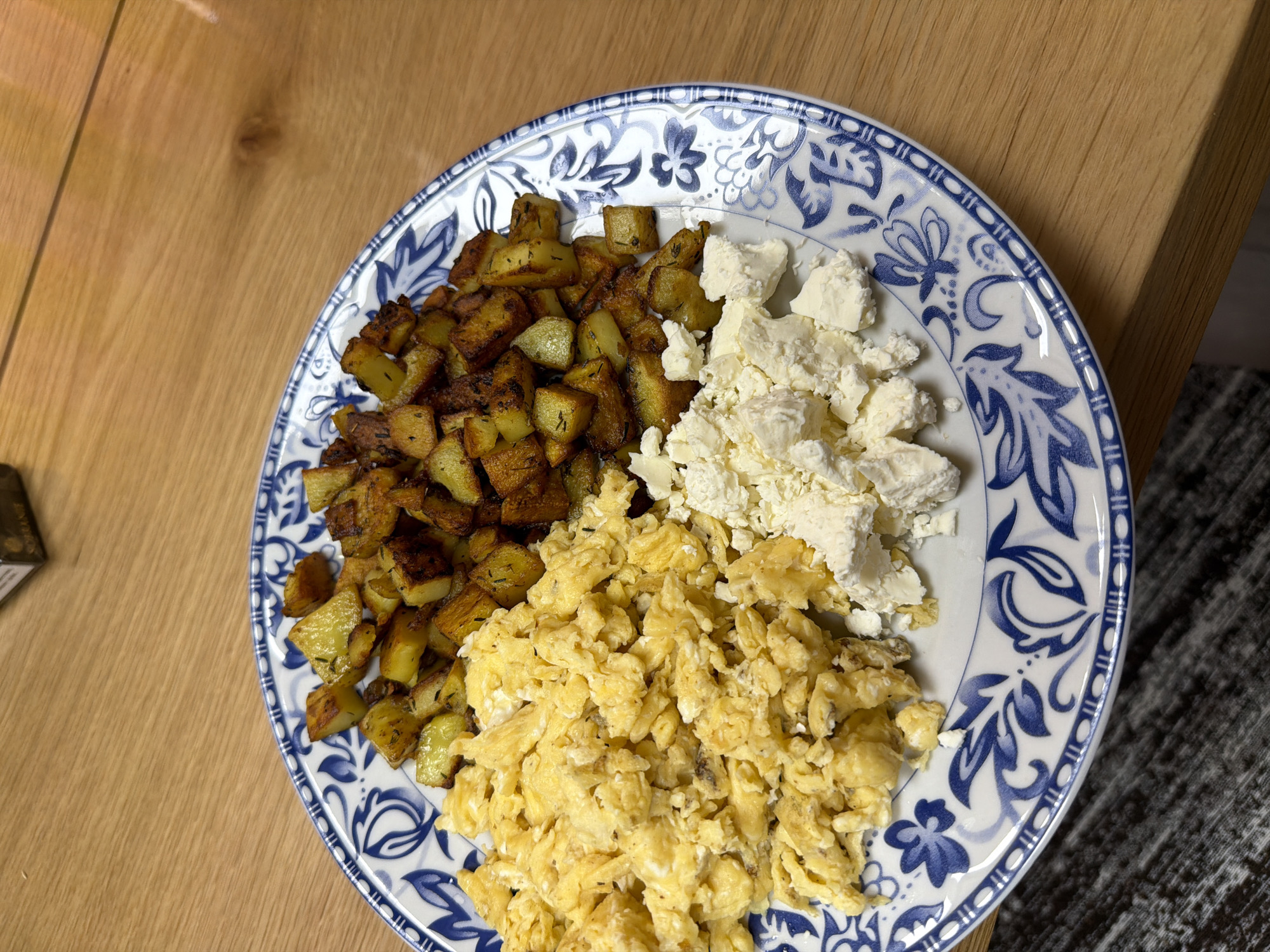 A top-down view shows a plate of food on a wooden surface. The plate, adorned with a blue and white floral pattern, contains three distinct portions: diced, roasted potatoes, scrambled eggs, and crumbled white cheese, likely feta. The potatoes exhibit varying degrees of browning, some darker than others. The scrambled eggs appear soft and lightly cooked. The crumbled cheese is presented in irregular clumps. A small, rectangular object, possibly a package or container, is visible in the extreme lower left corner. The wooden surface has a natural, light-brown tone.
The image was not taken in Panagyurishte, Bulgaria.