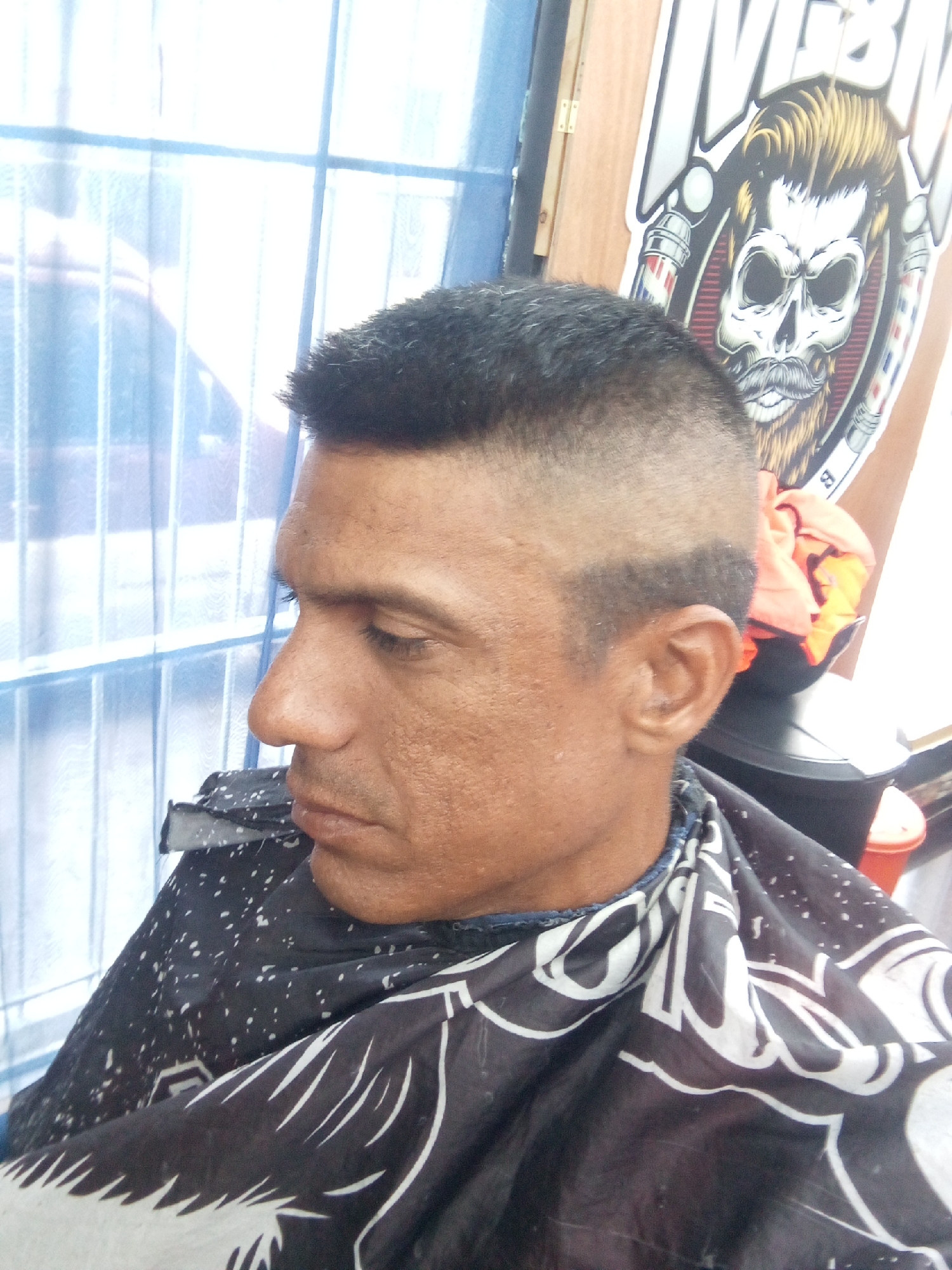 A medium shot depicts a man seated in a barber chair, his head turned to the left. He is wearing a black and white barber cape. The man has short, dark hair, styled in a fade haircut. The background includes a window with blue trim, a wooden panel with a barber shop logo featuring a skull, and various barber shop equipment. This image was most likely taken in Juan Griego, Venezuela.