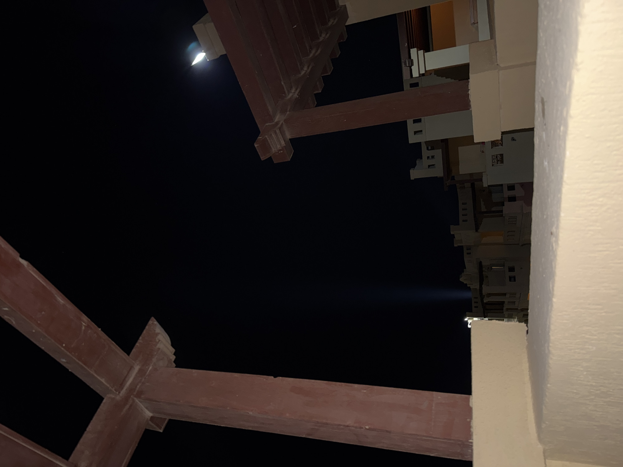 A low-angle, medium shot captures an outdoor night scene with a dark sky and architectural elements. In the foreground, a textured, light-colored wall runs horizontally across the bottom of the frame. To the left, a thick, reddish-brown, square pillar extends upwards, with a horizontal beam of the same color intersecting it. To the right, a portion of a reddish-brown pergola structure with visible beams and supports is visible. In the middle ground, a line of buildings with a muted color palette is illuminated from within, suggesting interior lighting. The buildings have a multi-story design with visible windows and balconies. A vertical beam of light emanates from a light fixture on one of the buildings, extending upwards into the dark sky. A faint crescent moon or light source is visible in the upper right quadrant of the sky, possibly a street light or lamp on a rooftop. The scene appears to be situated in an urban or resort setting.