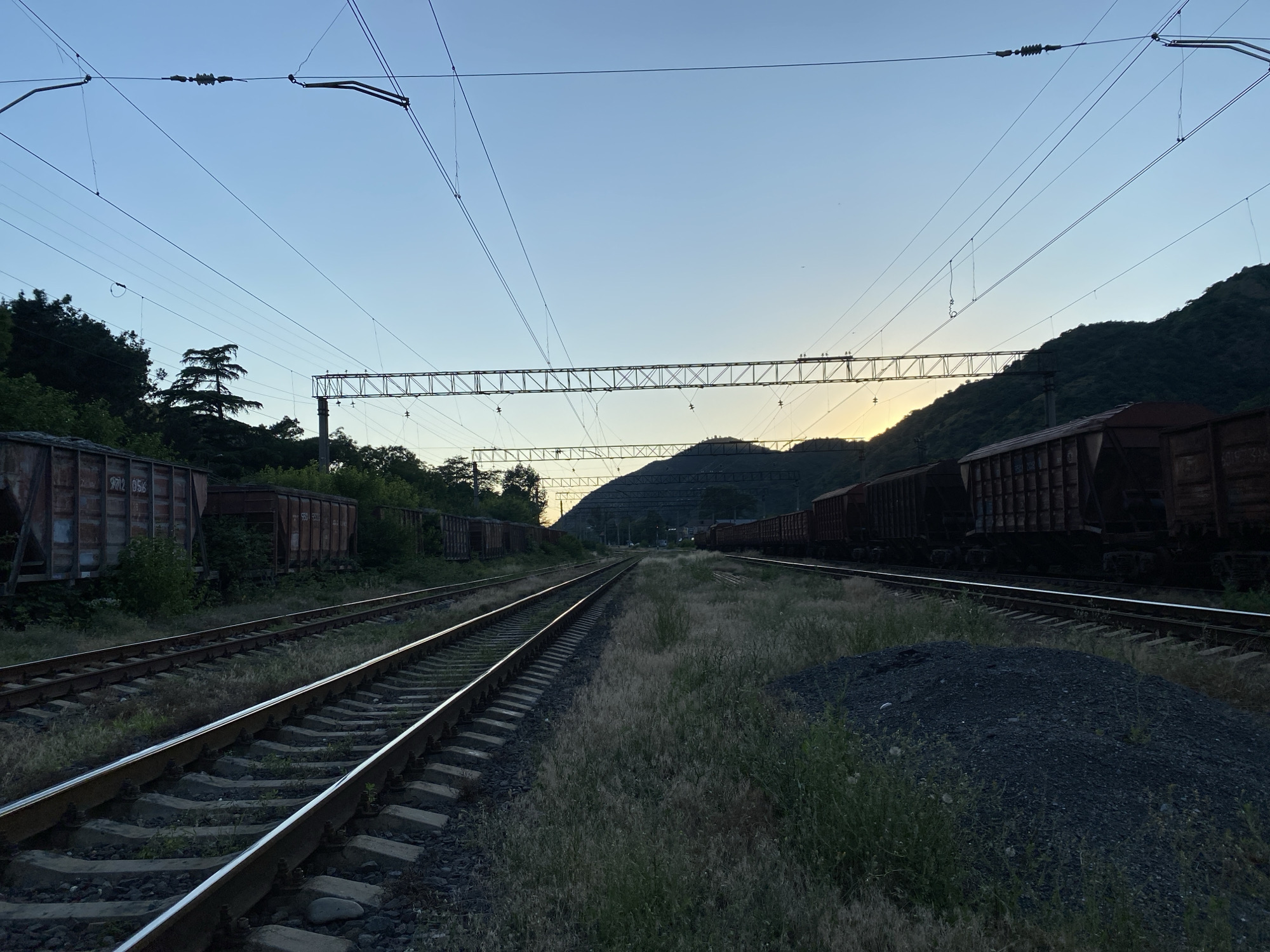 The image depicts a railway yard with multiple tracks running parallel to each other, extending into the distance. On either side of the tracks are parked freight train cars, many of which appear to be empty. The landscape is characterized by rolling hills and mountains in the background, with dense vegetation lining the sides of the tracks. Overhead, a complex network of power lines and support structures crisscrosses the sky, indicative of an electrified railway system. The sky is a clear blue, transitioning to a soft yellow near the horizon where the sun is setting, casting a warm glow over the scene. The foreground shows a pile of dark, granular material, possibly coal or gravel, situated beside the tracks. The overall impression is one of an active, though perhaps dormant at this moment, industrial railway area.
