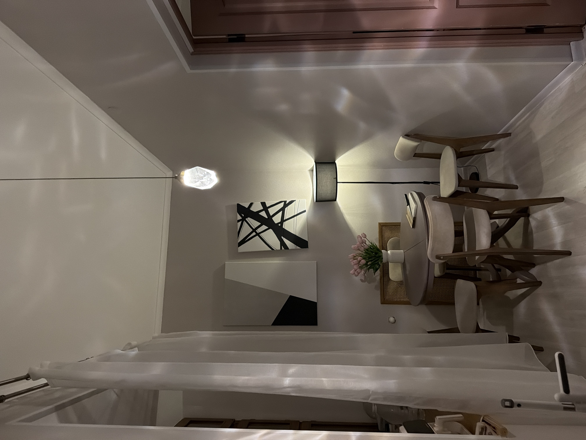 The image depicts an interior scene, likely a dining nook or a small seating area within an apartment. The room is dimly lit, with warm light emanating from a floor lamp and a hanging pendant light fixture with a crystalline bulb. The walls are painted a neutral, light grey color.
On the left side of the image, a sheer white curtain hangs from a rod, partially obscuring a vertical wooden structure and a white door handle with a vertical bar. This suggests the area might be near an entrance or a partitioned space.
In the center of the image, a round, dark-colored dining table is set with placemats and what appear to be a notebook and pen. The table is surrounded by three chairs. Two chairs are fully visible, upholstered in a light beige fabric with wooden frames. One chair is partially obscured by the table. Behind the table, a wicker-backed chair is visible. A white vase containing a bouquet of pink tulips sits on the table.
Above the table and chairs, two abstract paintings hang on the wall. The painting on the left is divided into geometric shapes of grey and black. The painting on the right features black lines on a white background.
To the right of the paintings, a tall, black floor lamp with a dark grey cylindrical shade casts light and shadows on the wall. Further to the right, a pink-toned door with a vertical dark handle is visible, ajar, revealing a sliver of another room.
The floor is light-colored wood. The overall impression is one of a cozy, modern, and minimalist interior.
This image is not identifiable as being in Moscow, Russia.