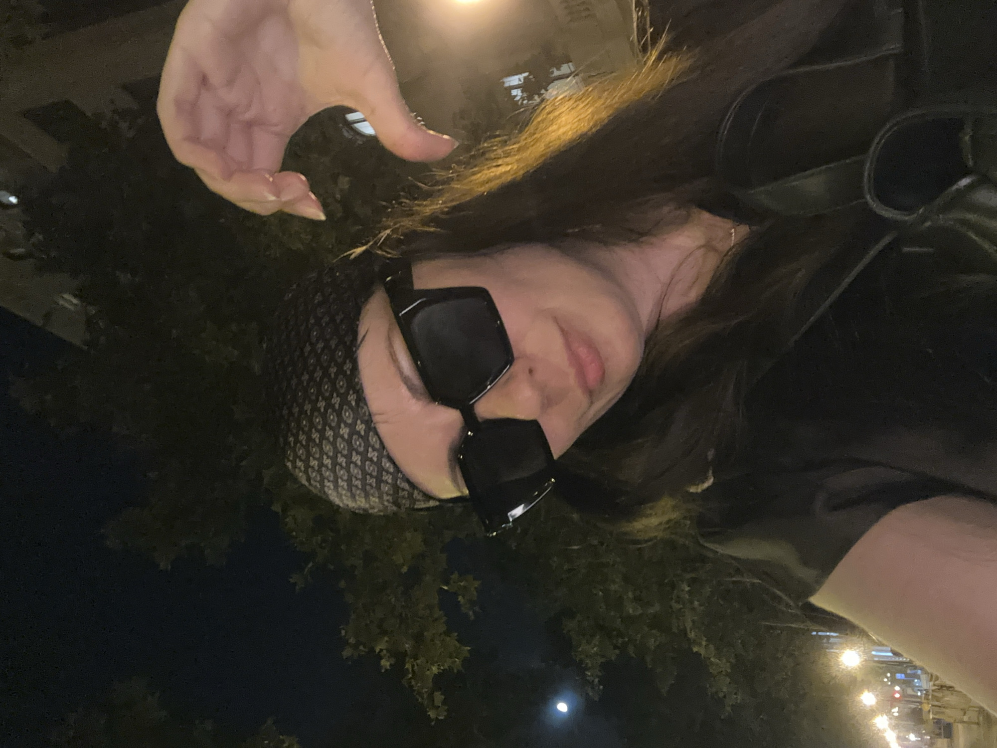 In this nighttime selfie, a woman with long, dark hair is pictured against a backdrop of trees and city lights. She is wearing large, square, black sunglasses and a patterned bandana. Her right hand is positioned to form a heart shape near her face. The scene suggests an outdoor urban setting, possibly a street or park in Barcelona, Spain, given the streetlights and architectural hints in the background, and the presence of a faint moon. The overall lighting is dim, with artificial lights creating warm glows.