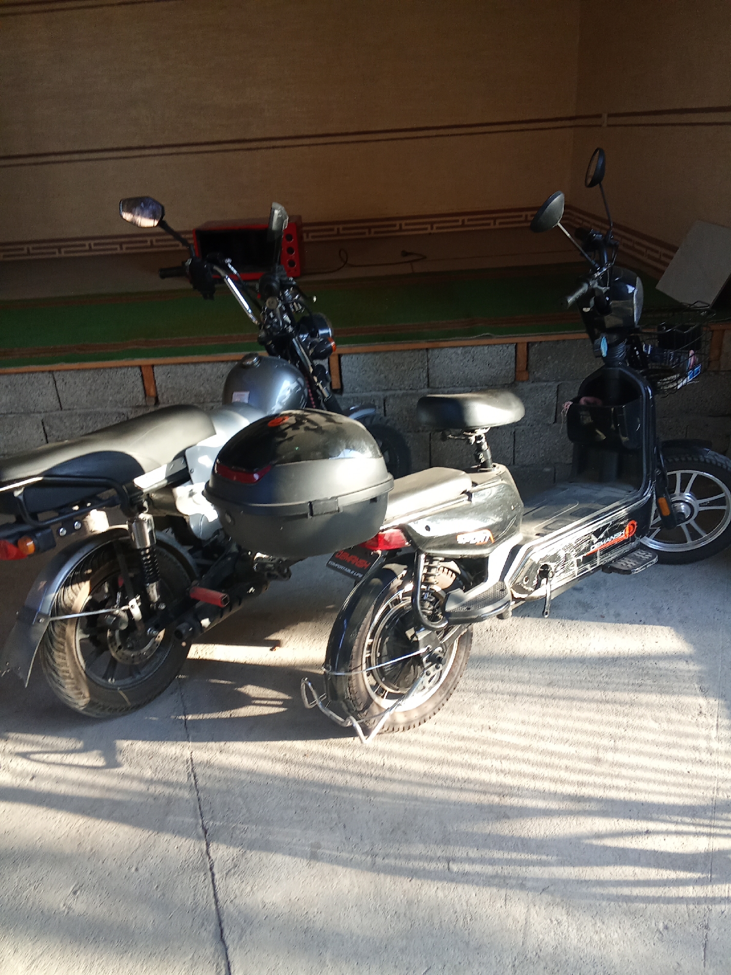 Two electric scooters are parked side-by-side in an outdoor setting, possibly a covered parking area or a sheltered walkway. The scooter on the left appears to be silver and black, with a large black storage box mounted behind the seat. The scooter on the right is primarily black, featuring a basket at the front and a similar storage box at the rear. Both scooters have visible tires, wheels, and handlebars.
The background consists of a textured wall with decorative horizontal bands, and a green surface that could be artificial turf or carpet, suggesting a recreational area or a public space. The ground beneath the scooters is a concrete surface with visible cracks and is partially covered in sunlight, casting distinct shadows that indicate it is likely daytime, with the sun at an angle. The lighting suggests it could be late afternoon or early morning. There are no people visible in the image.
On the right scooter, some text is partially visible on its side, reading "DEMANSHE". The overall scene is static, with the scooters simply parked. The environment appears to be a practical, somewhat utilitarian space. The location context provided is Fergana City, Uzbekistan.