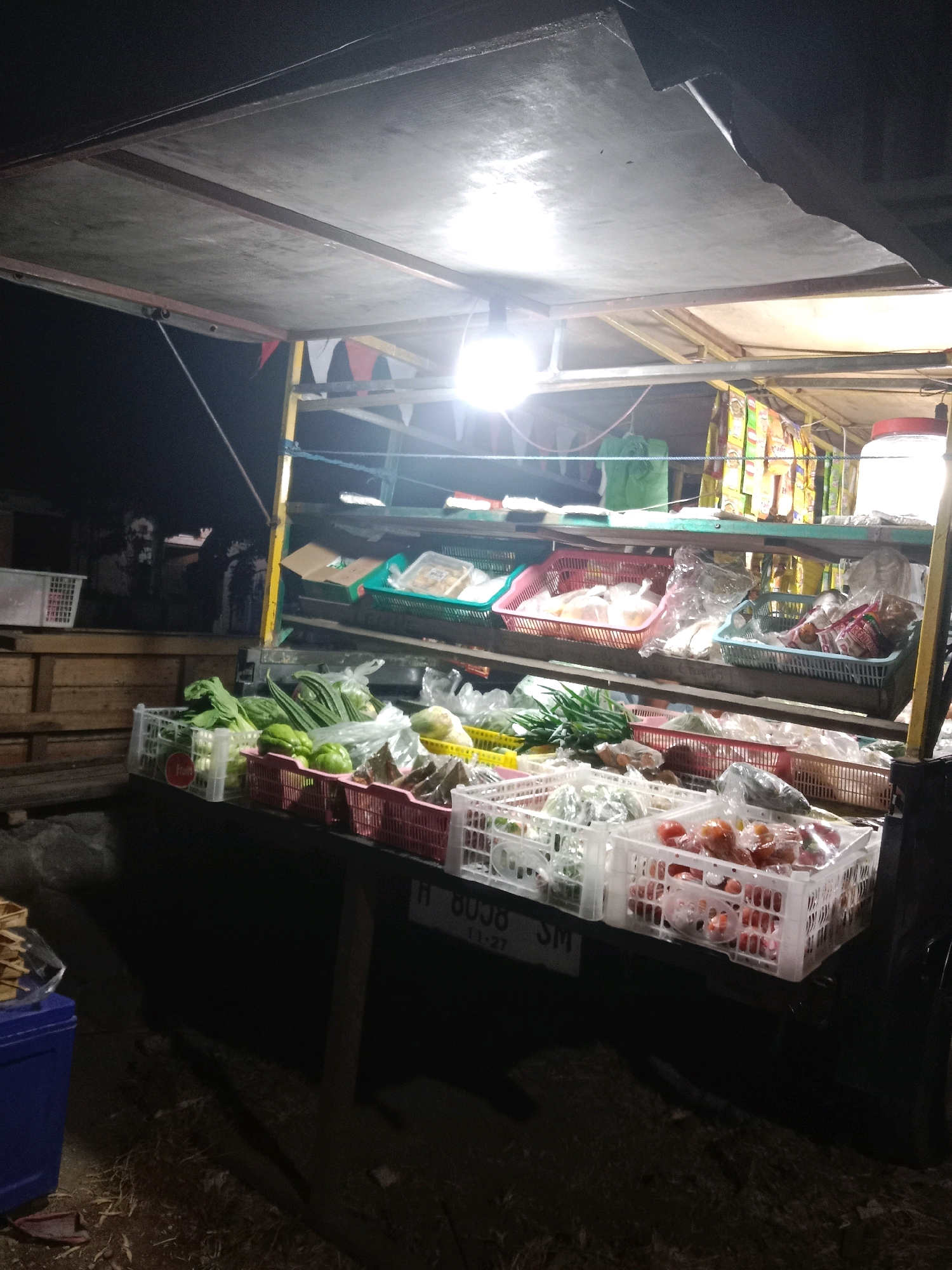 This image depicts an outdoor stall at night, likely in Langensari, Indonesia, selling fresh produce. The stall is illuminated by a bright overhead light, casting a strong glow on the goods. The main display features a variety of vegetables arranged in white and pink plastic crates. Visible produce includes green leafy vegetables, onions, gourds, and tomatoes. Some items are packaged in plastic bags. Above the produce, shelves are stocked with various packaged goods, some in colorful wrappers, possibly snacks or other dry items. A large, translucent container is visible on the top right shelf. The stall itself appears to be constructed with a metal frame and a corrugated roof. In the foreground, there is a black surface with a license plate that reads "H 8038 SM". The ground around the stall is dark and appears to be covered in dirt and straw. A blue plastic bin is partially visible on the left. The background is dark, suggesting it is nighttime.