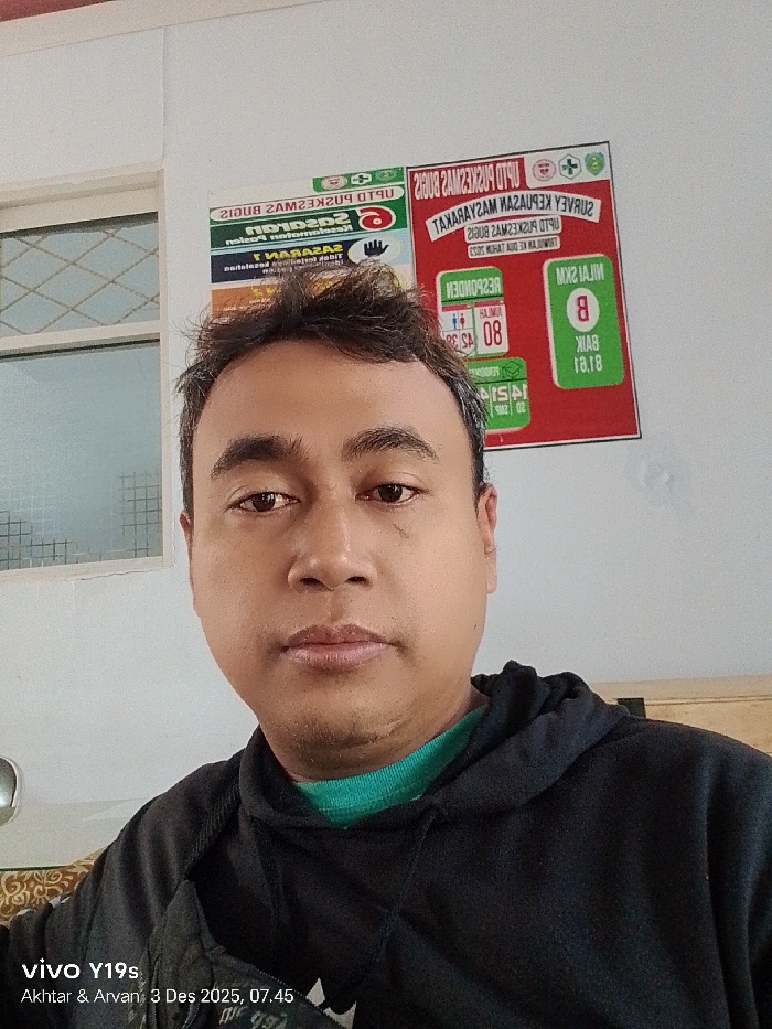 A person wearing a black hoodie with a green undershirt takes a selfie. Behind them are two posters with text and symbols, including a green section with the letter "B". A window with a metal lattice is visible to the left. The image contains a "vivo Y19s" watermark with the names "Akhtar & Arvan" and the date "3 Des 2025, 07.45". Some of the text from the posters is reversed and partially illegible.