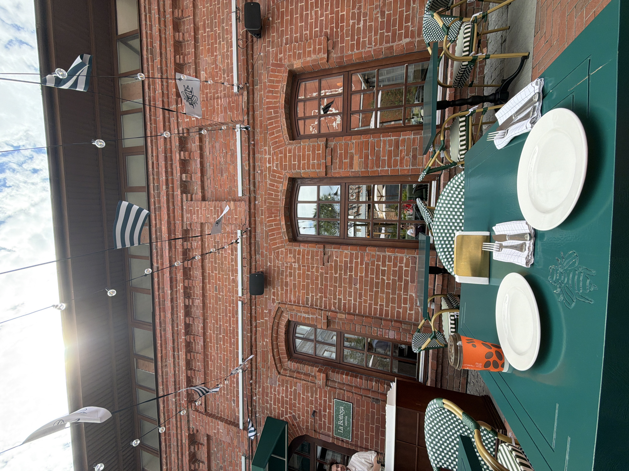 The image shows an outdoor seating area of a restaurant with red brick walls and a dark green table in the foreground. The table is set with two white plates, cutlery, and napkins. There is an orange to-go cup with a lid on the table. Behind the table, there are chairs with green and white patterned cushions and wicker frames. In the background, there are more tables and chairs, as well as large windows. Flags and string lights are hung above the seating area. A sign that reads "La Bottega osteria" is visible on the brick wall. There is a person standing behind a counter to the left of the frame. The scene appears to be in a city, possibly Ufa, Russia, given the context.