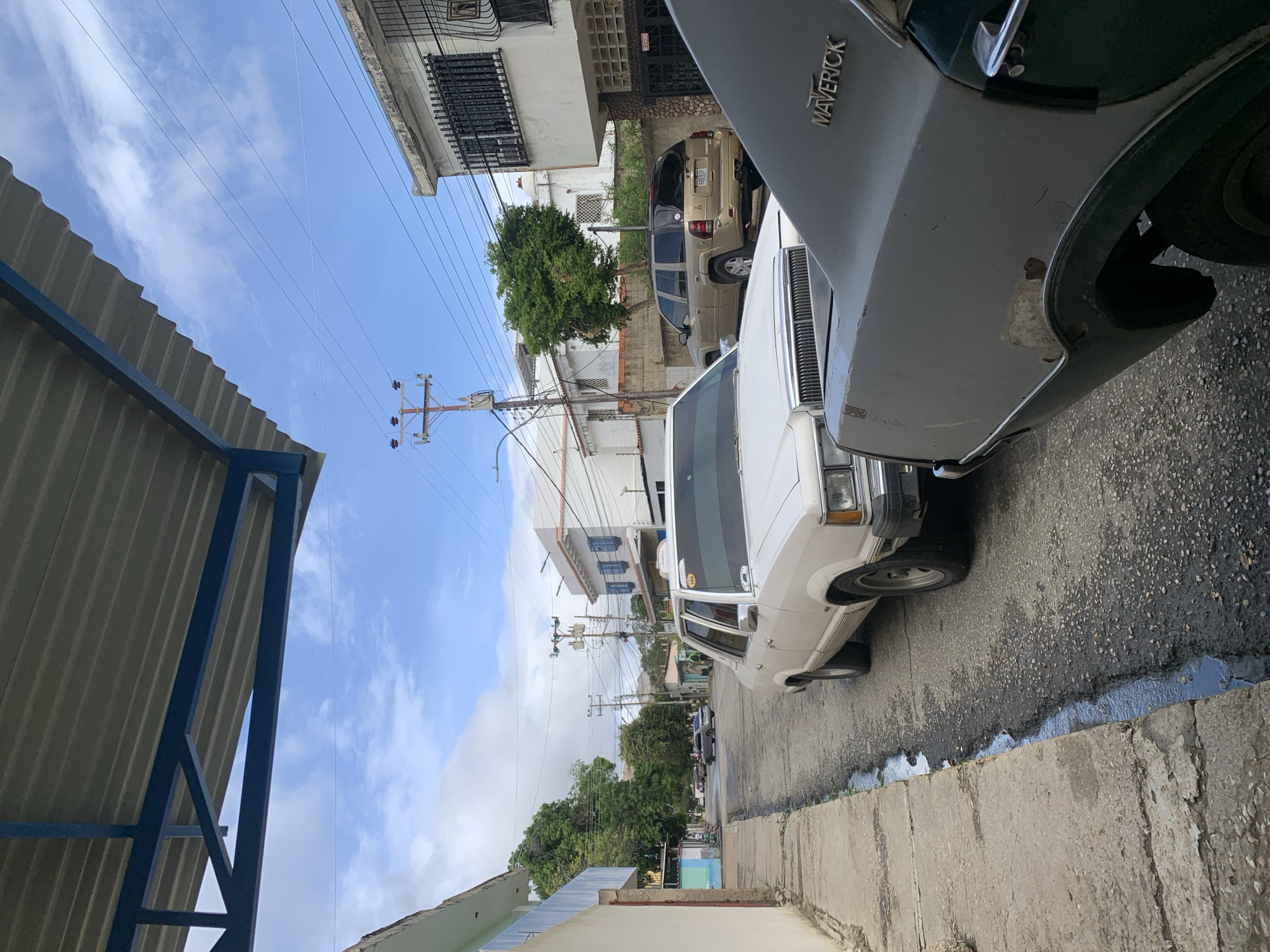 This image depicts a residential street scene in Santa Ana, Venezuela, under a bright, partly cloudy daytime sky. The perspective is tilted, with the street leading into the distance.
In the foreground on the right, a white Ford Maverick car is prominently parked, identifiable by the "MAVERICK" text on its rear quarter panel. The car shows signs of age and wear, including visible rust and paint damage on its side. The paved street alongside the vehicle is weathered, with a distinct channel or ditch containing standing water running along the curb.
Further down the street, a tan-colored SUV, possibly a Jeep Grand Cherokee, is parked on the right, with its license plate partially visible as "86B3P". Other cars are faintly seen further in the background. Buildings, likely residential, line both sides of the street, featuring various architectural styles, some with barred windows and balconies. A large tree with dense green foliage stands out mid-scene.
Utility poles heavily laden with power lines crisscross the sky, indicating a typical infrastructure for the area. On the far left, a structure with a corrugated metal roof and a blue metal frame is visible in the foreground. The weather is sunny with scattered white clouds. No people are visible, and no specific activity is taking place; the scene captures a moment of quiet daily life in the neighborhood.