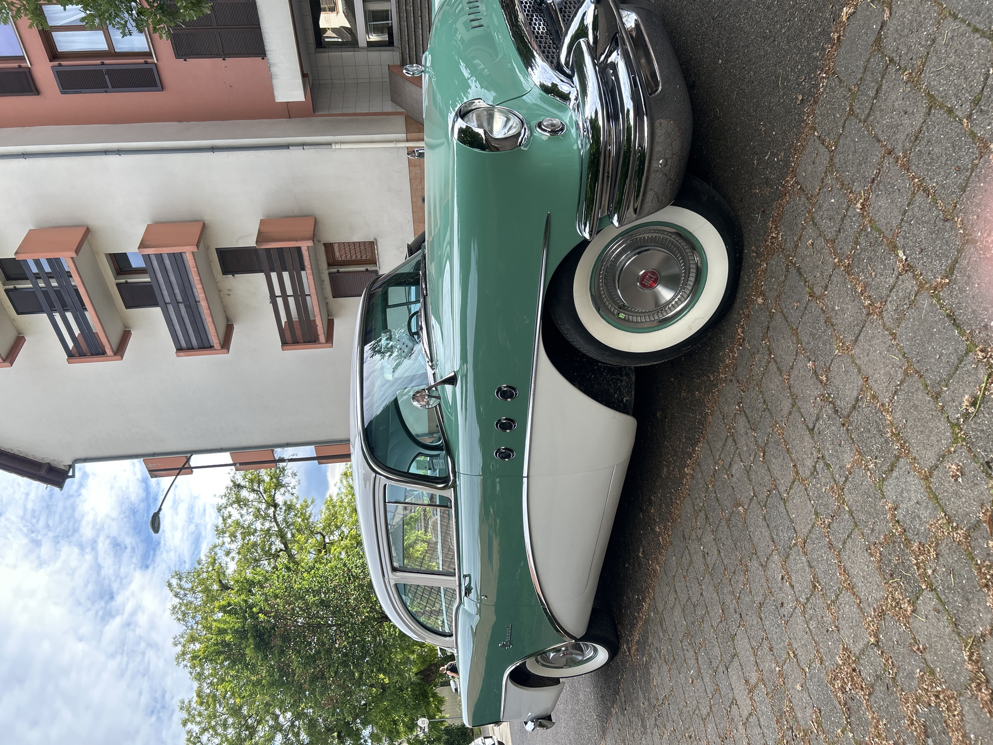 The image shows a vintage two-tone green and white Buick car parked on a brick-paved street. The car is positioned at an angle, showcasing its front and side. Its chrome detailing and whitewall tires are visible.
In the background is a light pink and white building with brown shutters and balconies, characteristic of European architecture. A green tree partially obscures a blue sky with white clouds.
The location is likely Strasbourg, France, given the architectural style of the building, which is common in the city. There are no people visible in the frame.