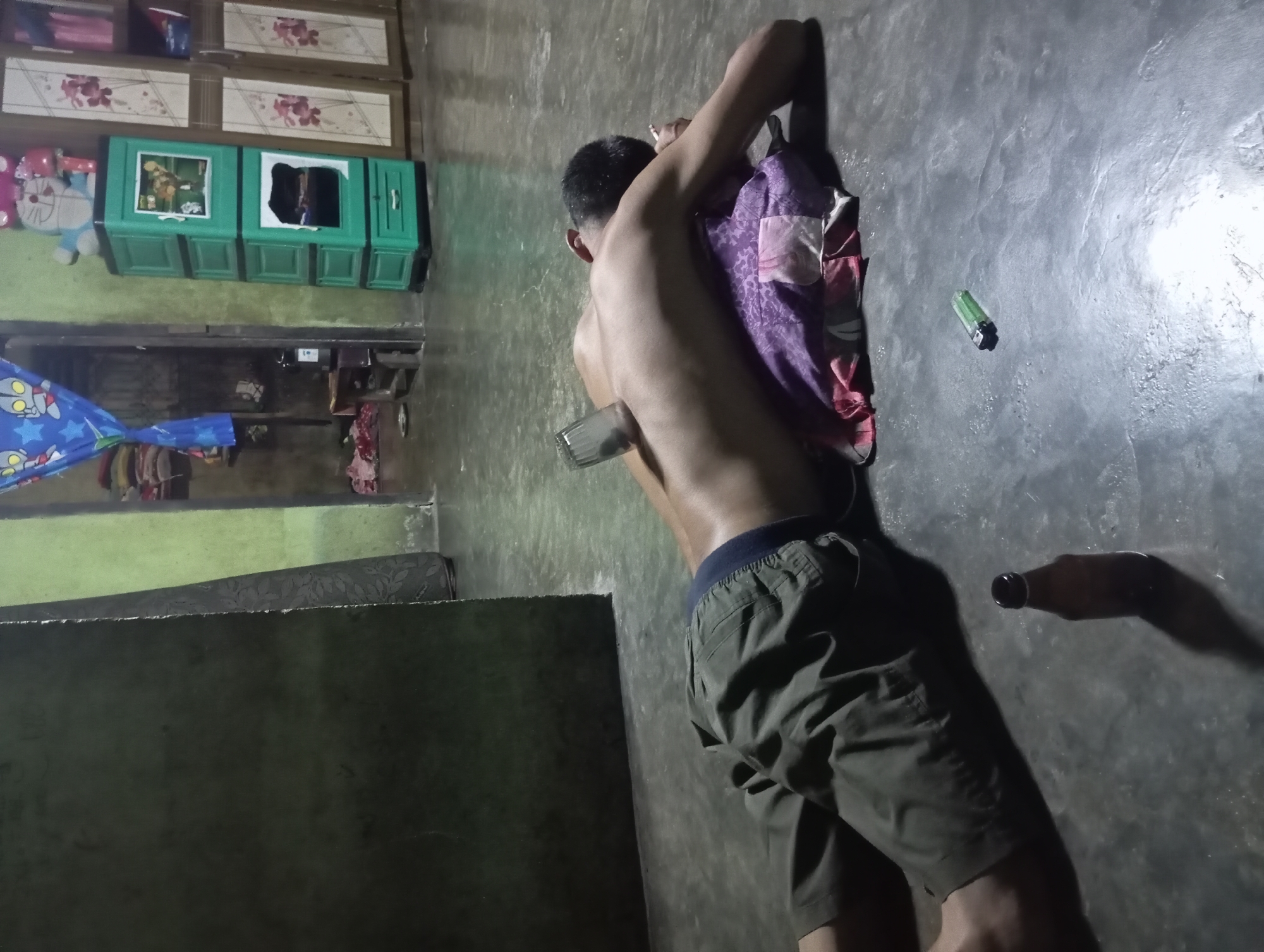 A shirtless man is lying face down on a concrete floor, with a glass object placed on his back and a pillow under his head. He is wearing green shorts. In front of him, a brown glass bottle stands upright, and a green lighter is on the floor. The scene appears to be indoors, with a green-painted wall in the background and a cabinet with drawers to the right. The overall impression is of a dimly lit room that might be a residential space in Rejosari, Indonesia.