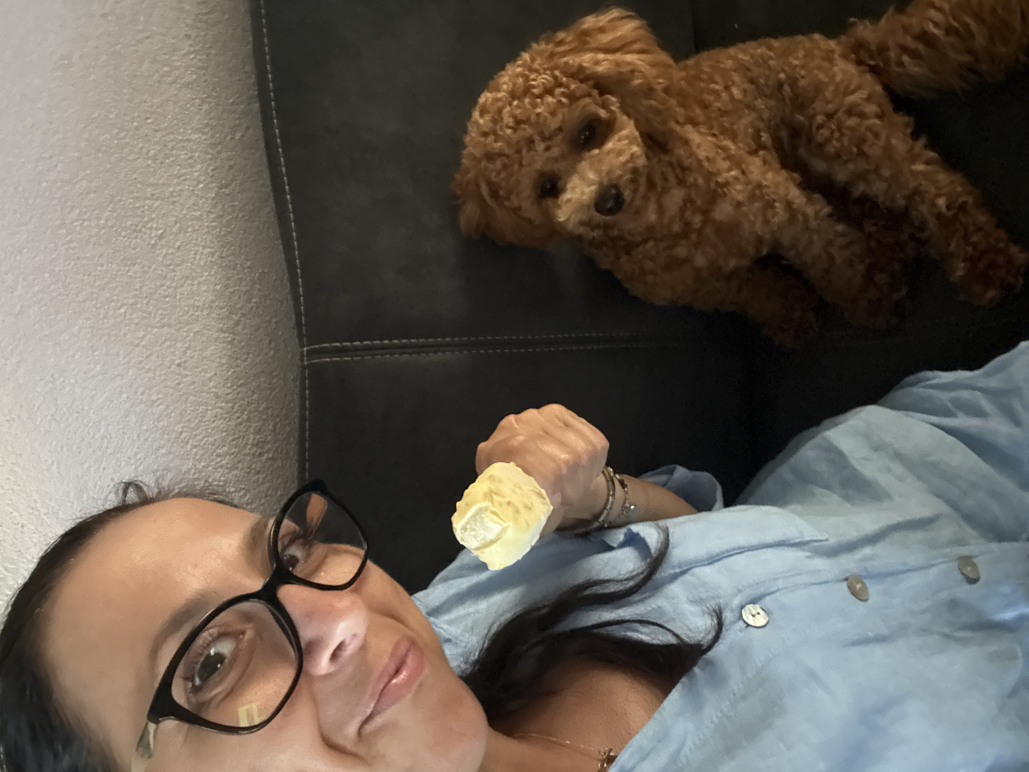 A woman and a toy poodle are sitting on a dark leather couch in what appears to be a living room. The woman is wearing a light blue button-down shirt and black-rimmed glasses. She is holding a piece of food, possibly a cookie or pastry, in her right hand and smiling at the camera. The poodle is a reddish-brown color and is sitting to the right of the woman, looking towards the camera with its head tilted. The background is a light-colored textured wall. The couch has visible stitching along the edges. The scene is dimly lit, suggesting it might be indoors with natural light coming from a window or artificial lighting. There is no clear indication of the specific location within Marum, Netherlands, but the setting suggests a residential environment.