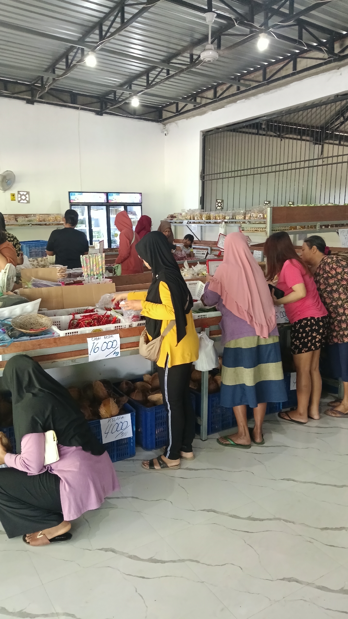 A medium shot captures the interior of a market stall in Pule, Indonesia, where several individuals are browsing and purchasing produce. The stall is organized with wooden shelves displaying various items, including red chilies, coconuts in blue crates, and other bagged goods. A white sign with handwritten text, reading "CABAT BESAR 16.000 KG," indicates the price of a specific type of chili. Another sign, partially visible, shows "KELAPA 4.000 BUJI" for coconuts.
Several women are the primary subjects. One woman in a yellow top and black hijab is examining chilies. Another woman in a pink hijab and a blue and yellow striped skirt is also looking at the produce. A woman in a pink t-shirt and a floral patterned top is standing to the right, seemingly engaged in conversation with another woman. A woman in a black hijab and a purple top is seated in the foreground, to the left. In the background, a man in a black shirt is visible near a refrigerator, and other shoppers are present. The market has a corrugated metal roof supported by a metal frame. Lighting fixtures are suspended from the ceiling, and a ceiling fan is visible. The floor is tiled with a marble pattern. The overall scene depicts a busy marketplace environment.