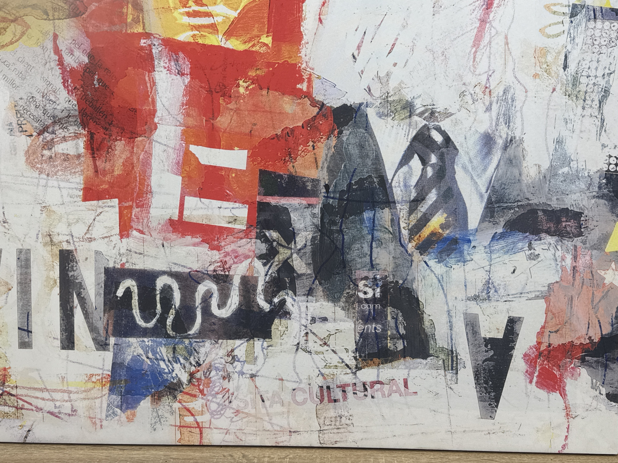 The image shows an abstract collage artwork with layers of paint, printed text, and graphic elements. Predominant colors are red, black, white, and gray. Large letters "N", "E", "W", "V", and "IN" are visible, integrated into the composition. Text fragments in various languages are incorporated. The style suggests an urban aesthetic, with distressed textures and overlapping forms. The artwork appears to be a physical object, possibly a canvas or board. The lower edge of the artwork is visible, resting on a wooden surface. No location details or people are discernible from the image.