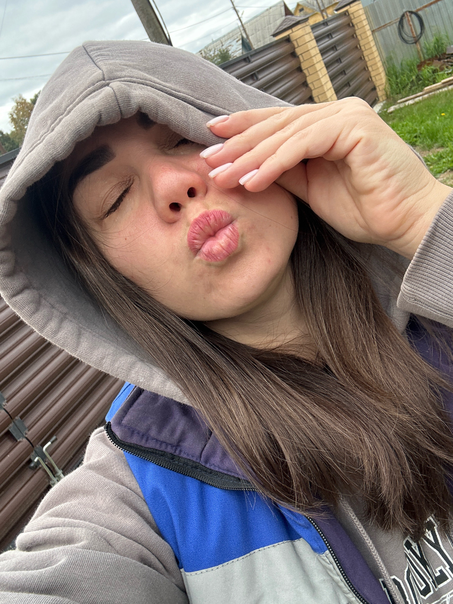A young woman, wearing a grey hooded sweatshirt and a blue and purple vest, is taking a selfie outdoors. She is puckering her lips as if to blow a kiss and has her right hand covering her right eye. Her eyes are closed. The background shows a wooden and metal fence, some overgrown grass, and a cloudy sky. The image appears to have been taken in a residential or rural area. There is no specific indication of the city of Chaikovsky, Russia, in the image.
