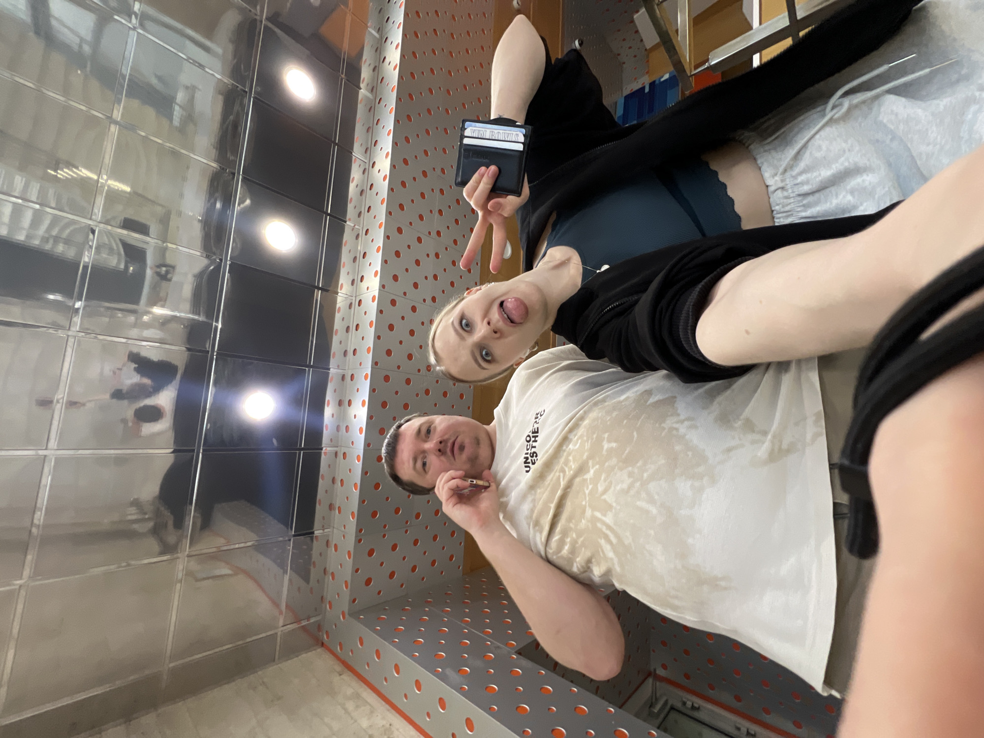 Two individuals are captured in a selfie within what appears to be an indoor facility, possibly a restroom, characterized by tiled walls and ceilings. One individual, a man, is holding what may be a cigarette or small object to his mouth. The other, a woman, is displaying a peace sign while sticking out her tongue and holding a wallet. The environment includes recessed lighting in the ceiling and perforated metal paneling with orange accents. The location is unspecified within the city of Khimki, Russia.