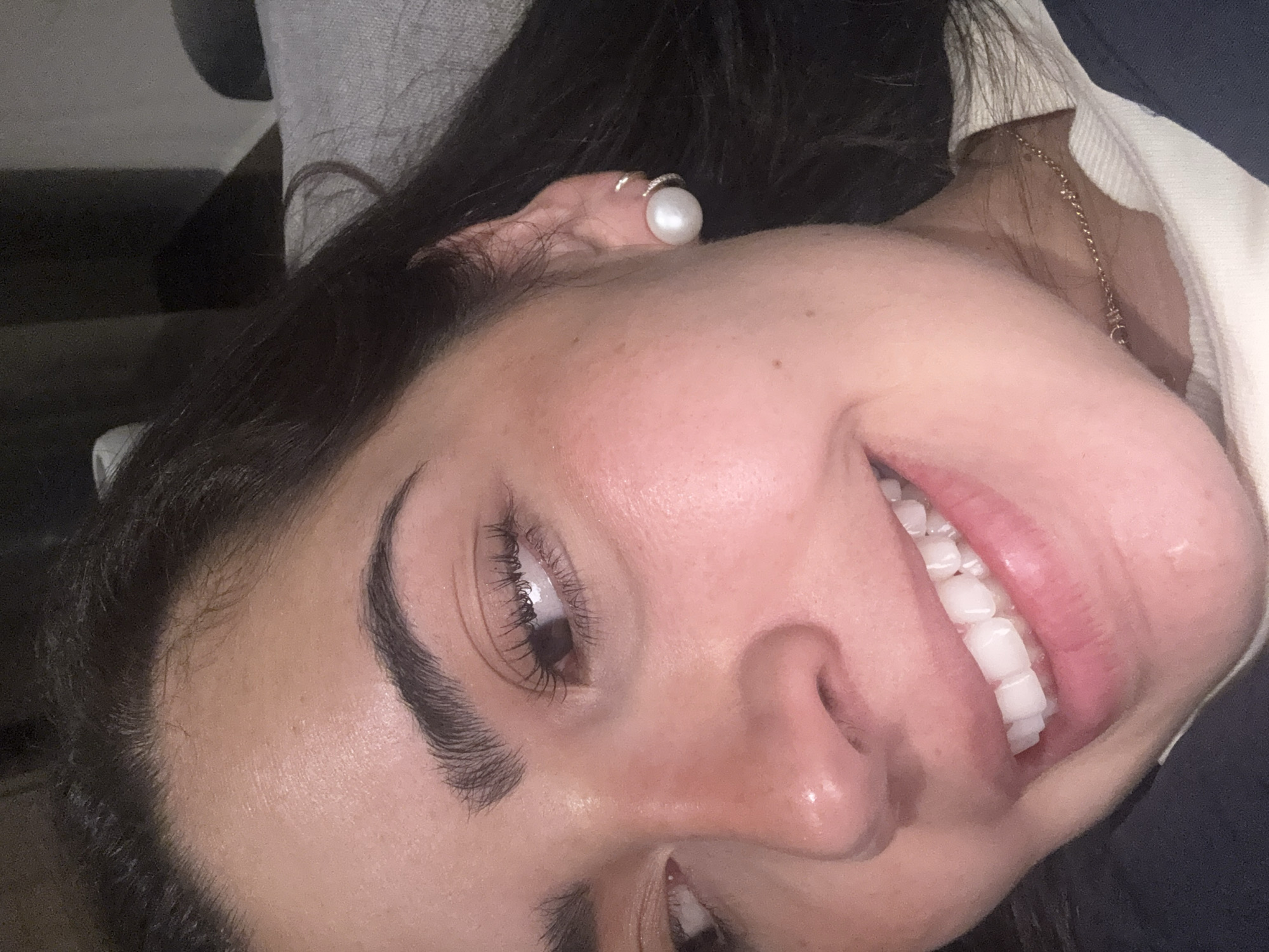 A close-up, tilted view focuses on a smiling woman. Her teeth are prominently displayed. The woman wears a large pearl earring. A fine gold chain is visible around her neck. No location information is discernible.
