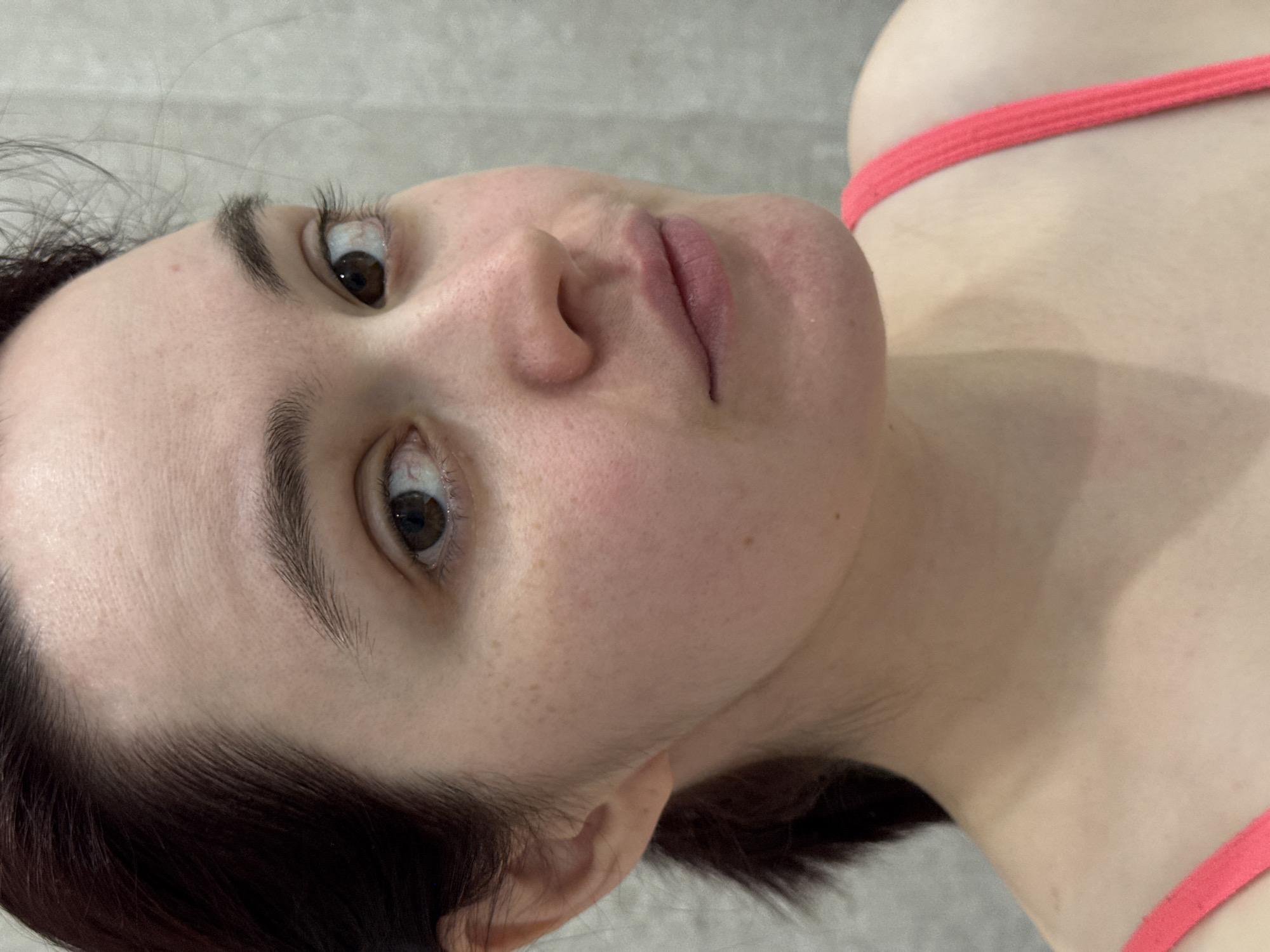 A close-up, medium shot shows a woman's face and upper torso. She is lying on her back, facing slightly upward and to the left. Her skin is fair, with freckles visible on her cheek. She has dark eyebrows and brown eyes. Her lips are closed. She is wearing a pink strap top. The background is a light grey. It is not possible to determine the location of the image, including whether it is in Izhevsk, Russia, from the visual information provided.