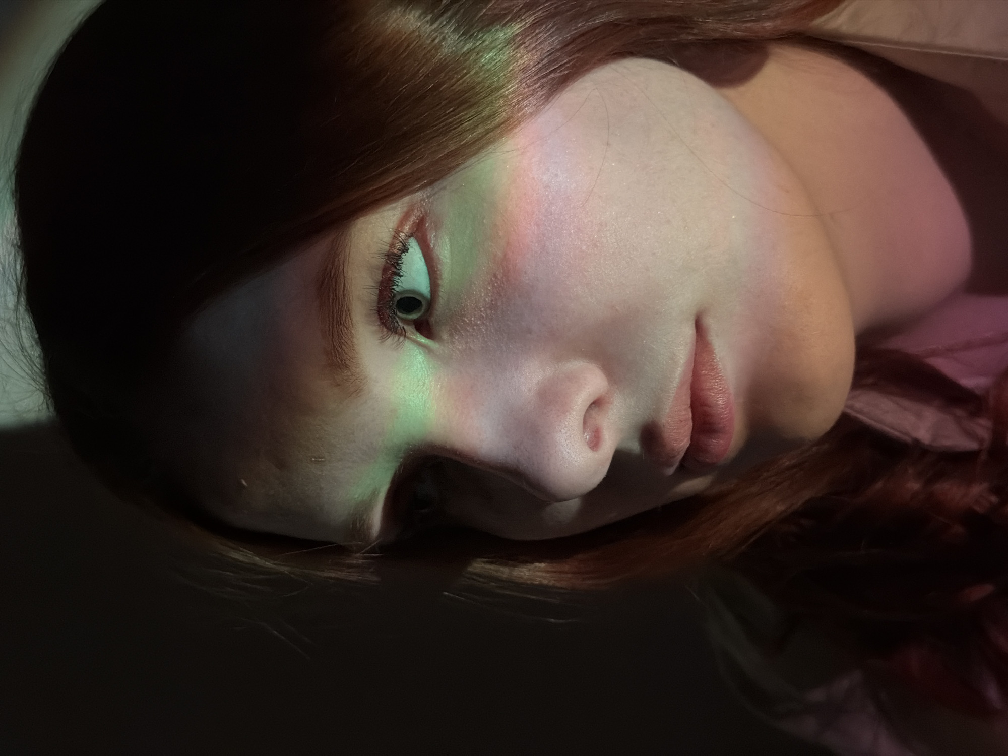 A close-up, artistic portrait features a person, likely female, with fair skin and auburn or reddish-brown hair, lying with their head tilted on a dark surface. The subject's eye is open and appears to be looking slightly upwards, while their lips are gently parted.
The most striking detail is a beam of colored light, possibly from a prism or projector, cast across the person's face. This light refracts into a spectrum of hues, predominantly greens and yellows, with hints of pink and purple, illuminating the eye, cheek, and part of the nose. The hair frames the face and blends into the surrounding darkness.
The overall scene is dimly lit, suggesting an indoor setting, with the colored light being the primary source of illumination for the facial features. The background is dark and indistinct, focusing attention entirely on the subject and the unique lighting effect. There is no visible activity beyond the person resting or posing. The expression conveyed is calm and contemplative. No text is visible in the image. The image provides no specific visual cues to its location, though it is stated to be Chaikovsky, Russia.