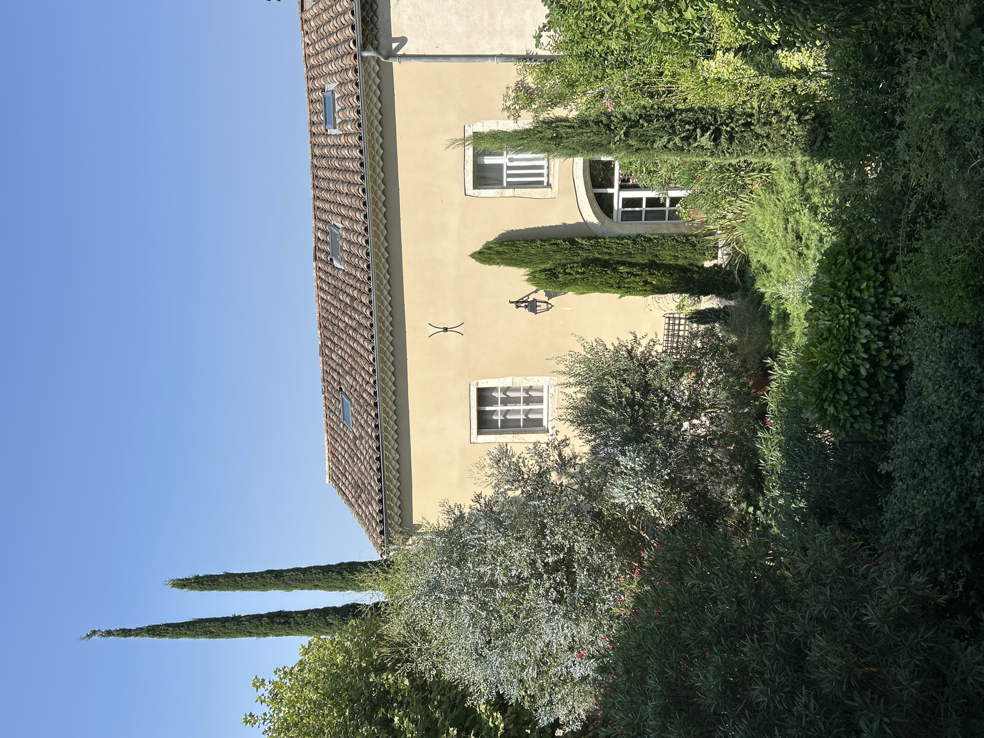 The image shows a two-story, light yellow house with a terracotta tile roof and two skylights. The house has several windows with white frames and panes, including a large arched window on the ground floor. A black lantern hangs on the wall to the left of the ground floor window. Tall, slender cypress trees stand in front of the house, along with various lush green trees and bushes in the foreground. The scene is bathed in bright sunlight, casting shadows on the foliage. The sky above is clear and blue. The location is Saint-Marcel-lès-Sauzet, France.