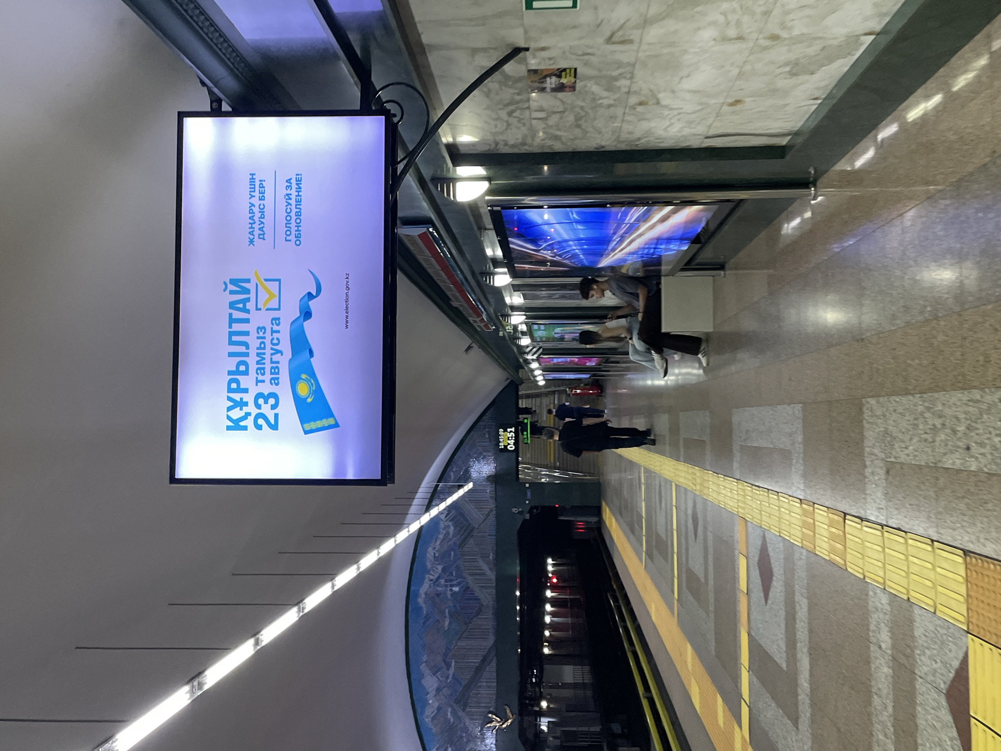 The image shows a platform in a subway station. A large digital billboard at the top of the frame displays an advertisement for elections in Kazakhstan. The billboard has text in Kazakh and Russian, a stylized image of the Kazakh flag, and the date "23 августа" (August 23). The website "www.election.gov.kz" is also visible. The subway station platform is clean and well-lit by fluorescent lights. Yellow tactile paving is installed along the edge of the platform for the visually impaired. Several people are present on the platform: a man in a black shirt and pants walks away from the camera, another man in a dark shirt and lighter pants sits on a bench, and a woman in a white top and jeans sits next to him. In the background, a train is visible on the tracks, and stairs lead up to another level of the station. A digital clock above the stairs displays "18:45:09" and "04:51". The walls of the station are decorated with a large mosaic depicting mountains. The overall impression is of a modern and functional public transportation space in Almaty, Kazakhstan.