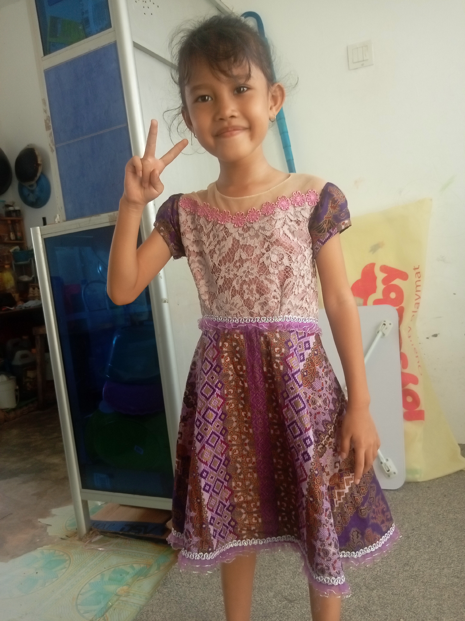 A young girl with dark hair and brown eyes, wearing a patterned purple dress, stands facing the camera and holds up two fingers in a peace sign. Her dress has a lace overlay on the bodice and a colorful, geometric pattern on the skirt. She is standing in an indoor environment, possibly a home, with a white wall behind her. To her left, a tall, reflective surface, likely a refrigerator or cooler, is visible. In the background, shelves with various items are partially obscured. To her right, a large, yellow plastic bag with red writing is propped against the wall. The floor appears to be tiled in some areas and carpeted in others. The overall lighting suggests it is daytime. The context of Batam, Indonesia, indicates this is likely a residential setting within the city.