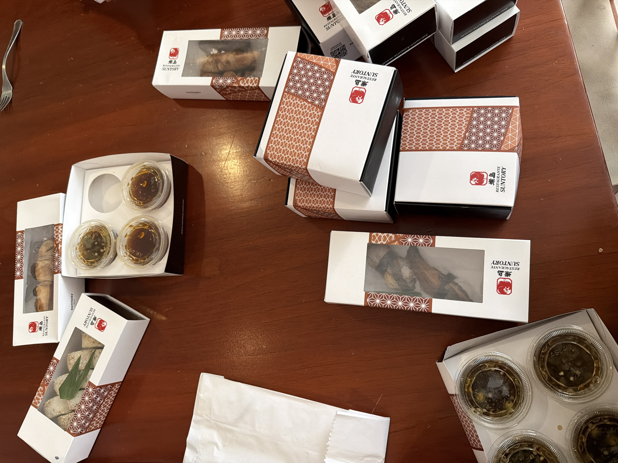 The image shows a variety of take-out containers from the restaurant Suntory, arranged on a brown wooden table. The containers are white with a brown geometric pattern and the restaurant's logo, which includes the name "RESTAURANTE SUNTORY" and the website "www.suntory.com.mx." Some containers have clear windows revealing the food inside, which includes sushi, tempura, and small sauce containers. There is also a white paper bag on the table and a fork in the upper-left corner.