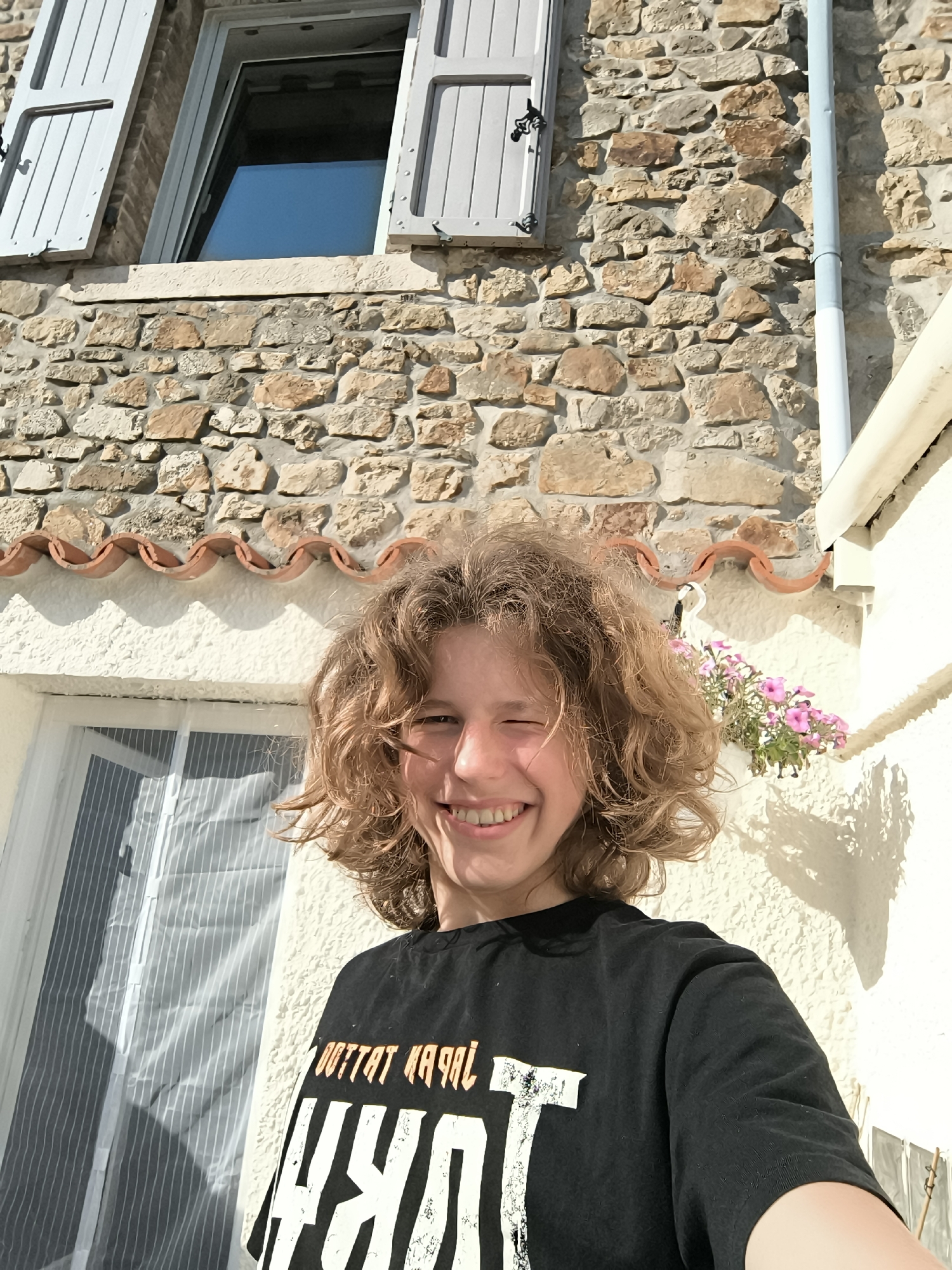 The image depicts a young person, likely a teenager, standing outside a stone building in Pélussin, France. The person has curly blonde hair, is wearing a black t-shirt with a white graphic design. The individual is smiling and winking at the camera. The background consists of a stone wall building with a window and light blue shutters. A white pipe runs vertically along the wall. There is a flower box with pink flowers hanging on the white wall. The building facade has a row of terracotta tiles. A window with a sheer curtain is visible to the left. The light suggests it is a sunny day.
