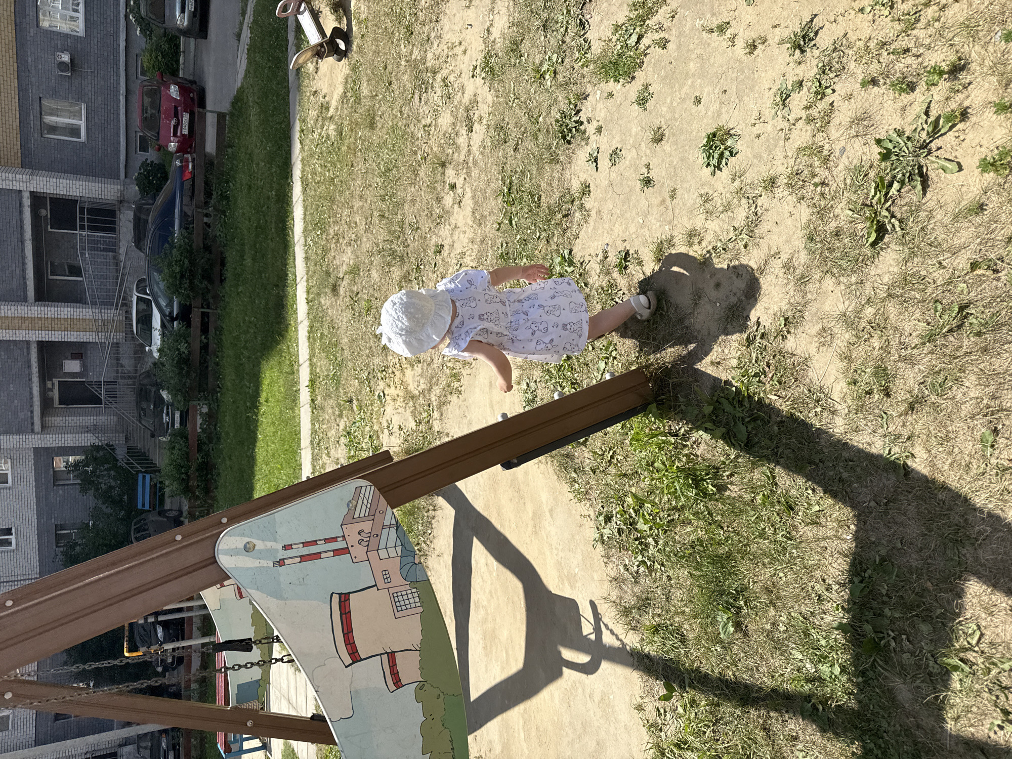 A child wearing a white sun hat and a white dress with a rabbit print walks away from the camera on a playground. The child is on a dirt and patchy grass area. To the left of the frame is a wooden swing set with a colorful panel depicting industrial smokestacks. A shadow of the swing set is cast on the ground. In the background are apartment buildings, parked cars, and a fence. The scene appears to be in a residential courtyard. The location is confirmed as Vladimir, Russia.