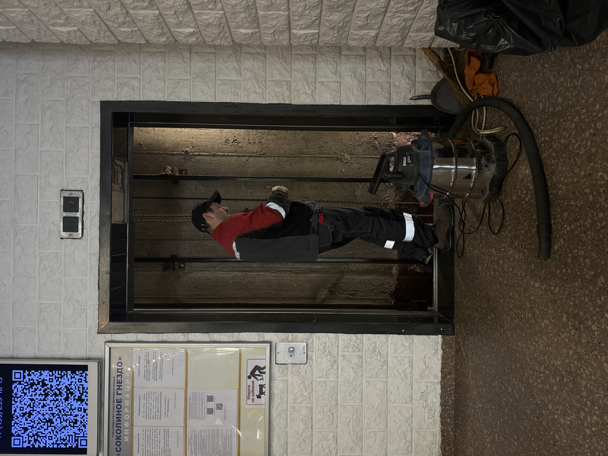 A maintenance worker is inside an open elevator shaft, apparently performing some sort of cleaning or repair. The shaft is lined with rough, wooden panels. The man is wearing a dark cap, a red and black jacket, and dark pants with white reflective stripes. A large silver and black shop vacuum cleaner with a long, flexible hose sits on the floor to the right of the shaft. To the left of the elevator opening is a white-tiled wall. A rectangular sign with a QR code, Russian text, and a phone number is visible. To the right is a framed information board with papers, as well as a small sign illustrating a person cleaning up after a dog, and an Otis elevator call button. A bag and other cleaning supplies are on the floor to the upper-right of the shaft.