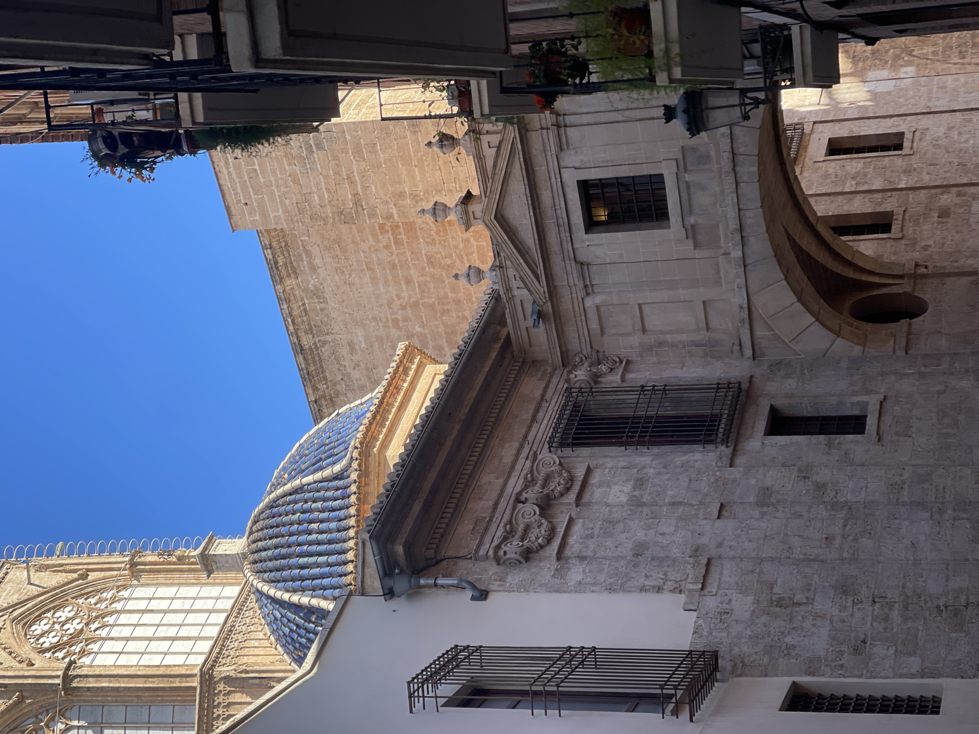 This is a low-angle, outdoor shot showing a building with a tiled dome and stone facade in València, Spain. The blue and white tiled dome is on the left side of the frame, with an adjacent building featuring Gothic windows and white facade elements. The stone building is a light tan color. There are several small, rectangular windows with metal grates. The top of the frame includes the bottom of a building. The sky is bright blue.