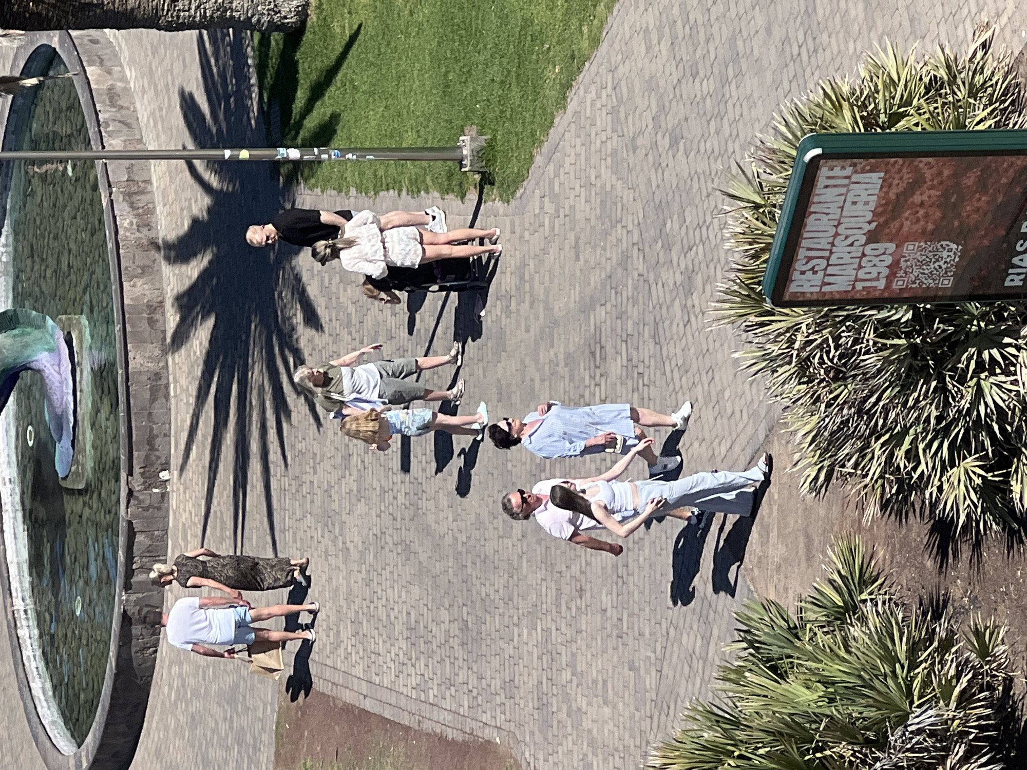 The image presents an elevated view of an outdoor public space, likely a plaza or park, in Maspalomas, Spain. There are multiple individuals walking along a stone-paved pathway. Some are in pairs, and some alone. The people are dressed in summer clothing. A circular fountain with a sculpture is a visible feature on the left side of the scene. A palm tree casts its shadow on the walkway. Vegetation is visible on the right side of the frame, including palm trees and signage that reads "RESTAURANTE MARISQUERIA 1989 RIAS B..." indicating a restaurant.